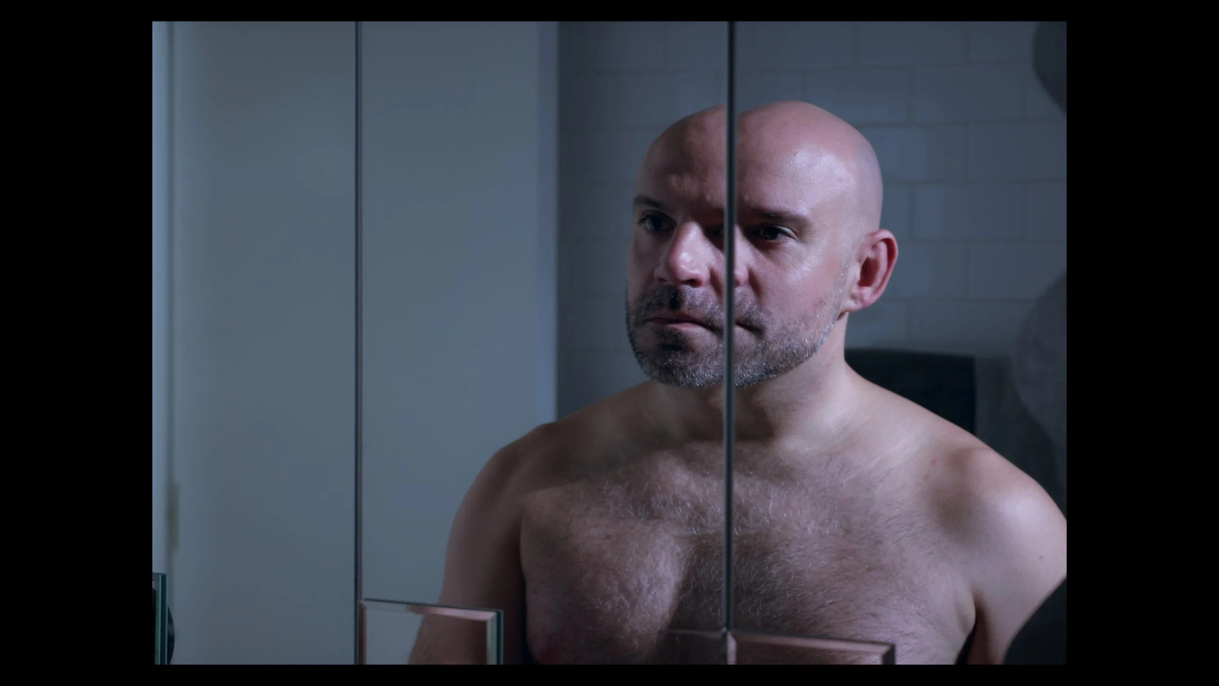 A shirtless bald man with a beard looks at himself in a mirror in a bathroom.