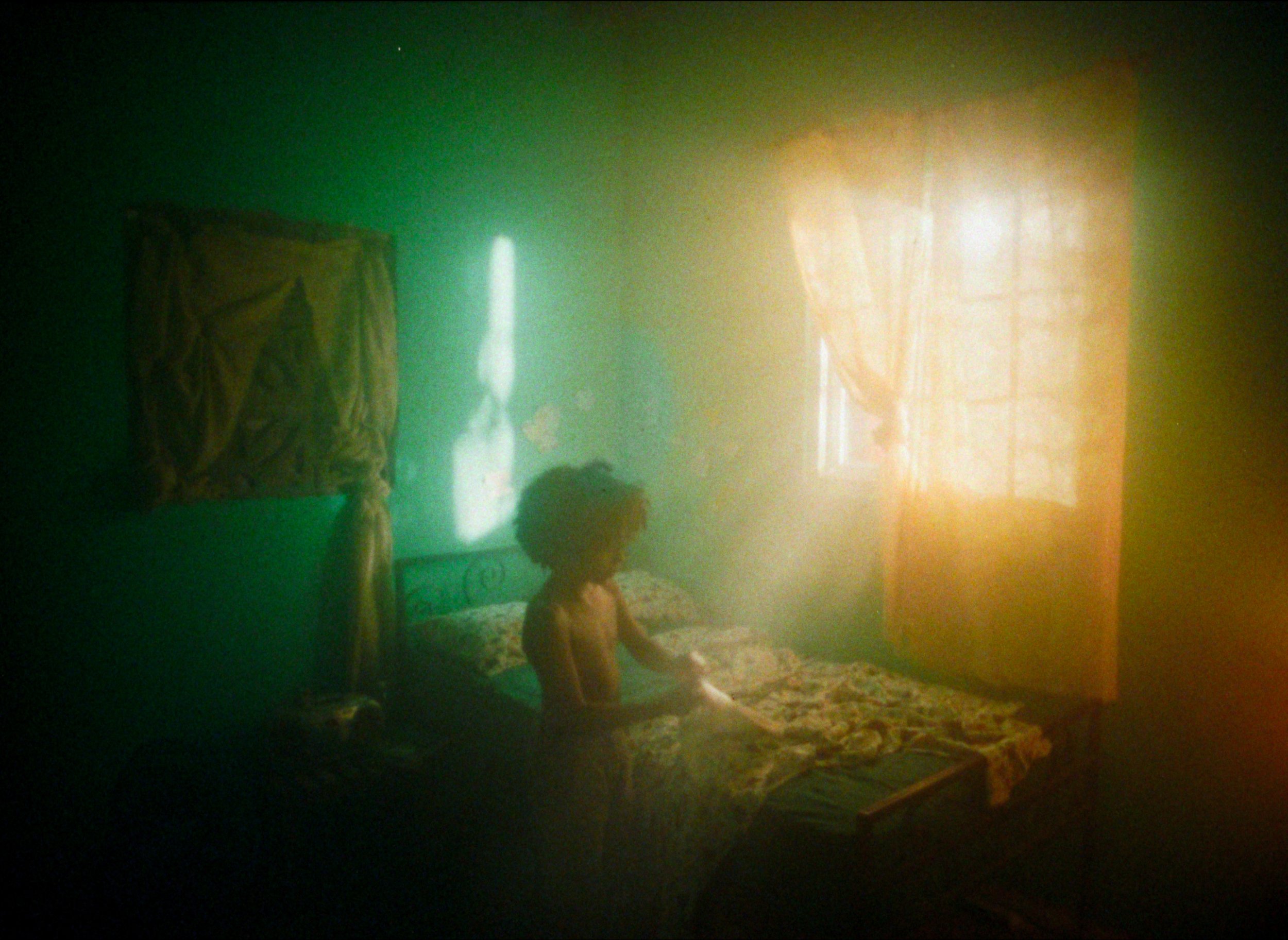 A young boy standing near a bed in a dimly lit room with green walls, a window with orange curtains, and sunlight filtering through the window.