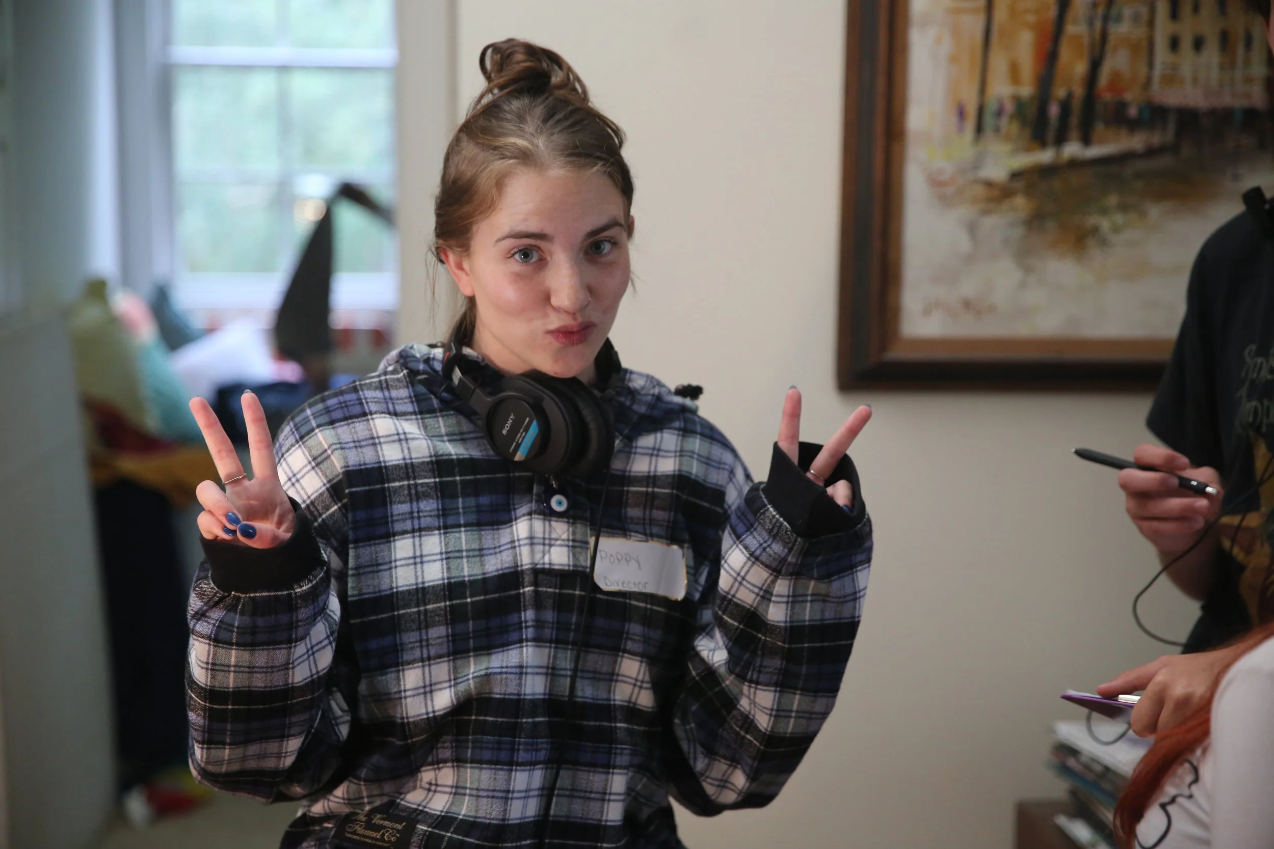 Young woman with brown hair in a messy bun, wearing a plaid shirt with a headshot tag labeled 'Poppy Director', making peace signs with both hands, standing indoors with colorful pillows and a window in the background.