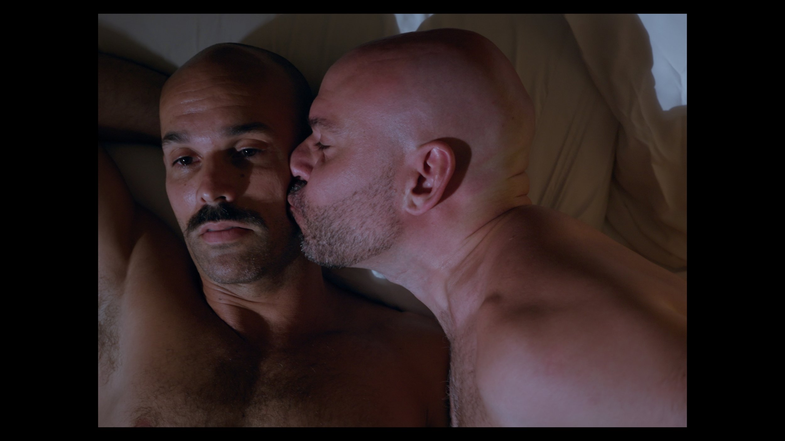 Two shirtless men lying close together on a bed, one kissing the other's cheek.