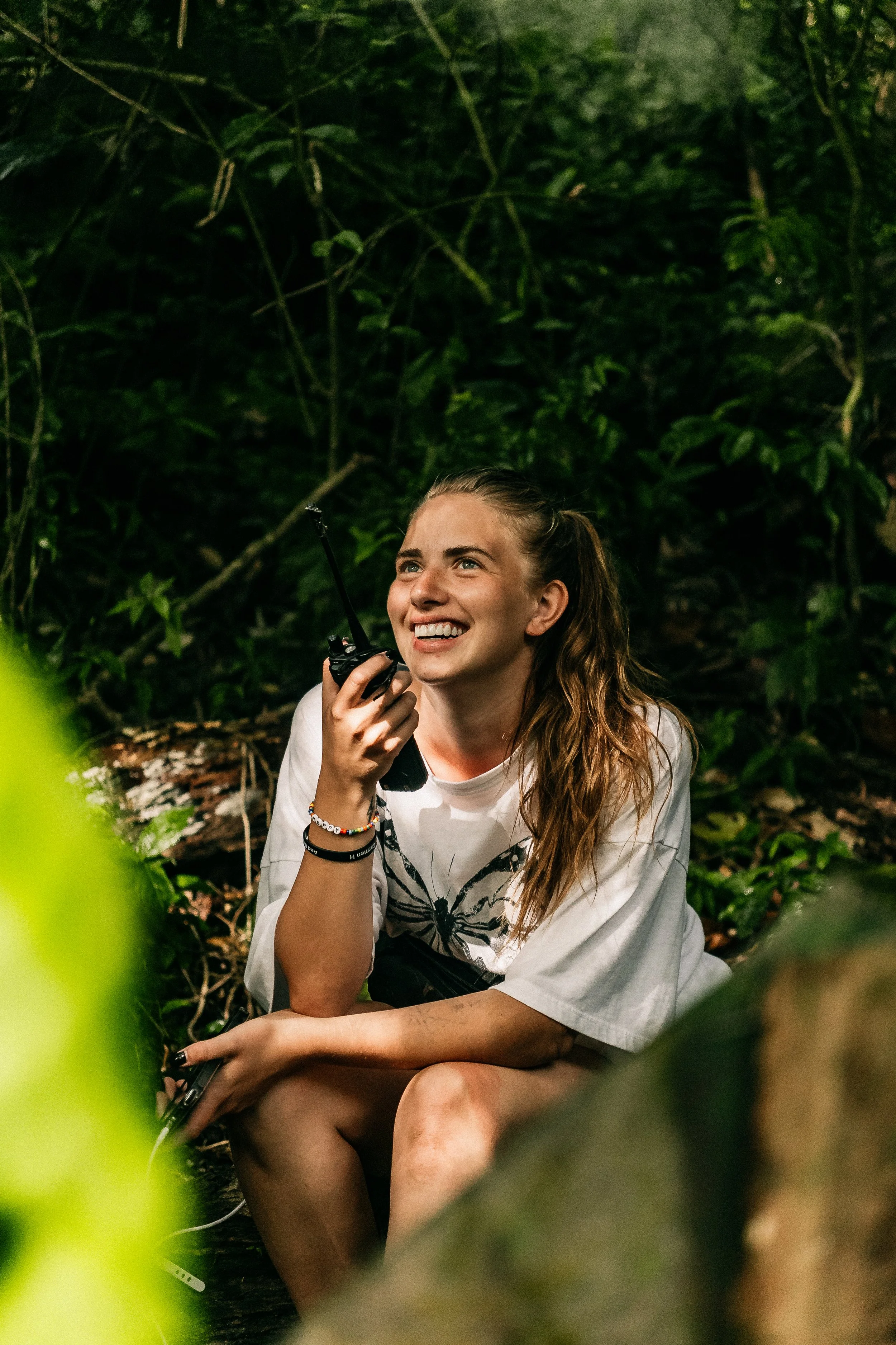 A young woman with long, wavy hair sitting in a lush green forest, smiling and holding a walkie-talkie.