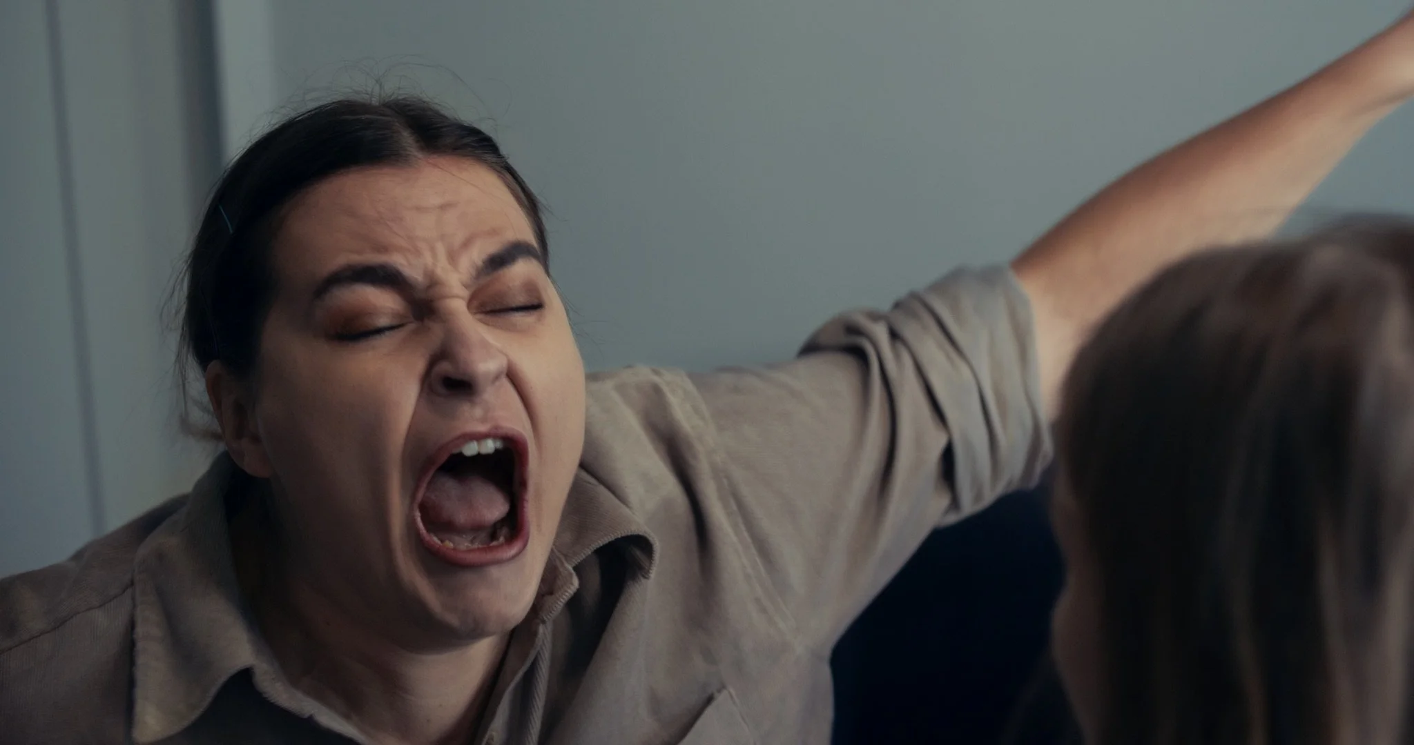 A woman with dark hair is yelling or screaming with her eyes closed and mouth wide open, reaching out with her arm.