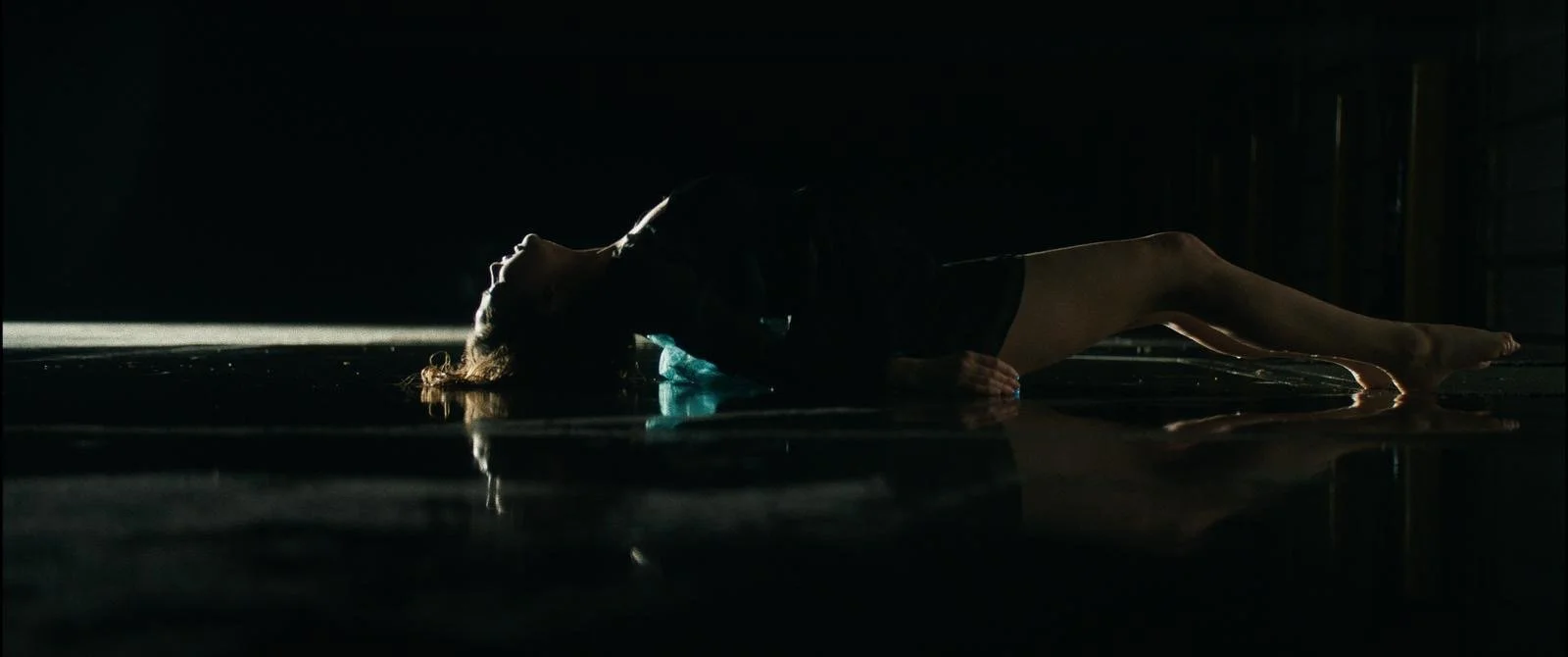 A woman lying on her back on a dark surface, with her head tilted back and illuminated by a light source from behind, creating a silhouette. She appears to be in a dimly lit room.