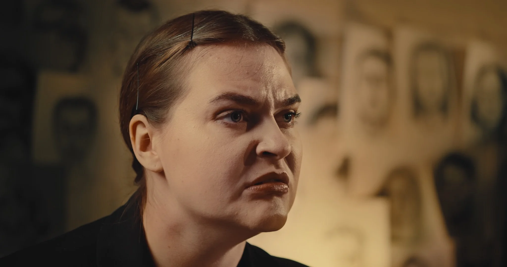 A young woman with her hair back, light makeup, and a nose ring appears to be upset or angry. The background features a blurred wall with drawn portraits of people.
