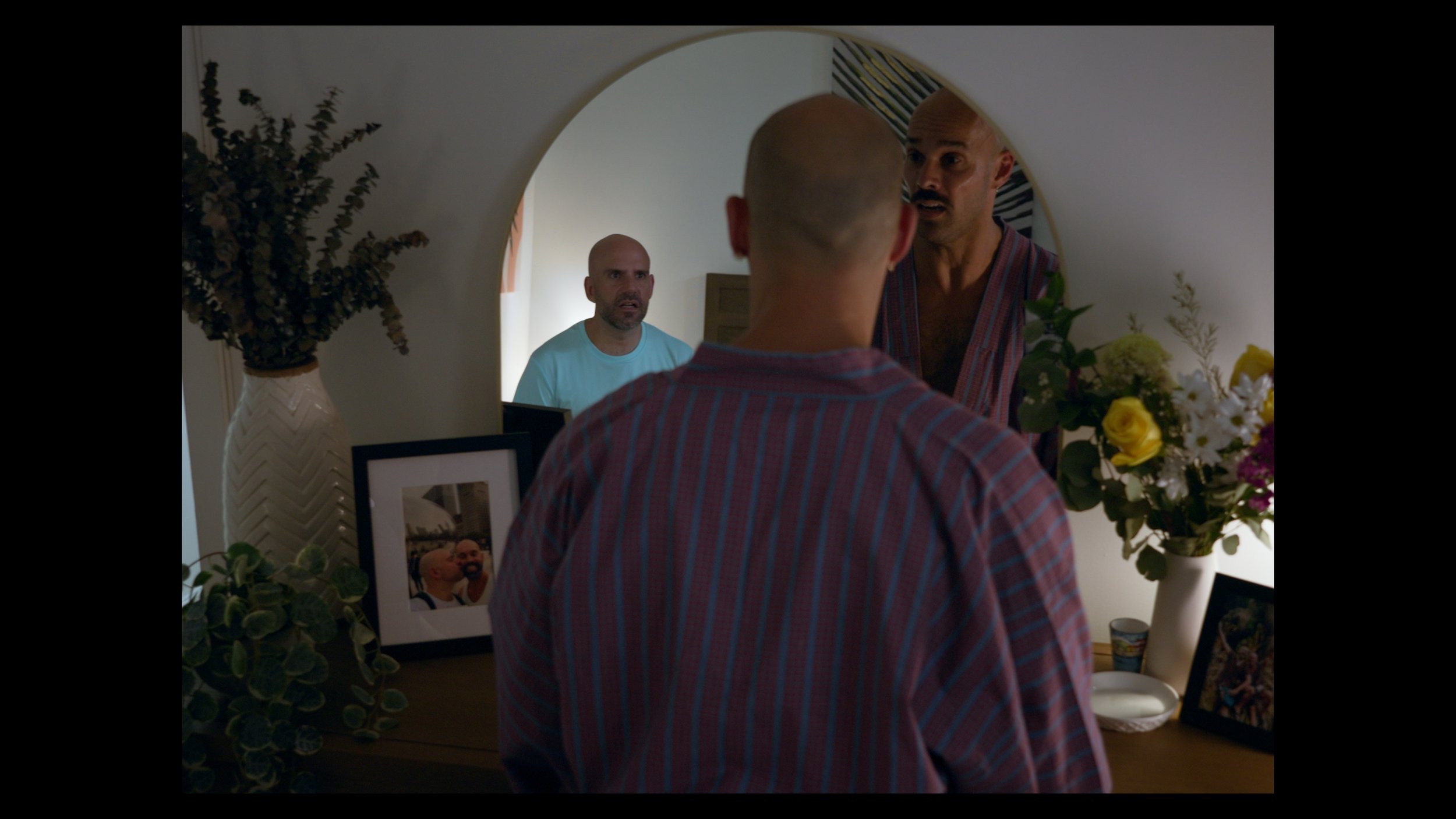 A man with a shaved head looking into a mirror, talking to another man who is also visible in the reflection, in a room with flower vases and framed photographs on a dresser.