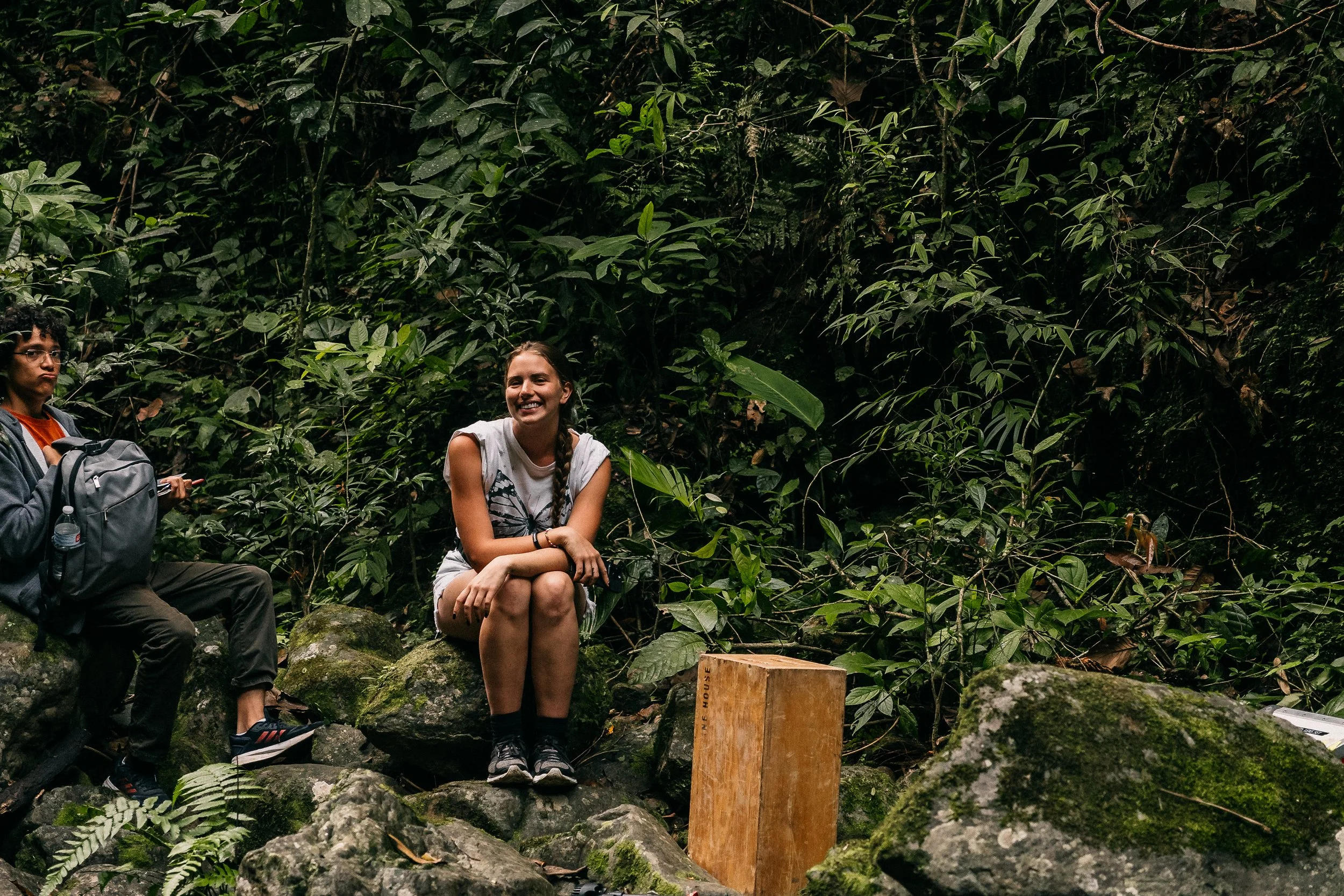 Two women sitting on rocks surrounded by lush green forest, one smiling and one looking away with a backpack.