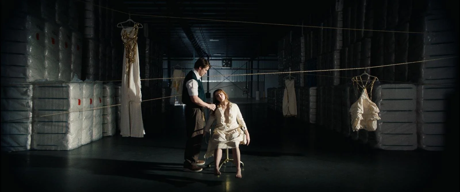 A woman in a white dress sitting on a stool in a dimly lit space surrounded by hanging dresses and stacks of large wrapped packages, with a man standing beside her.