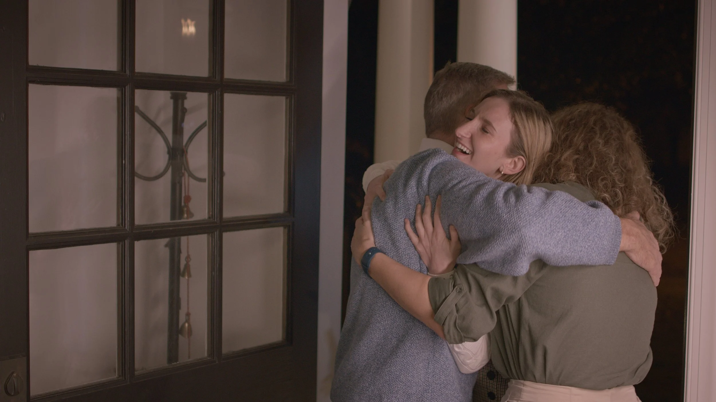 Three people hugging and smiling at night at a doorway, celebrating or greeting.