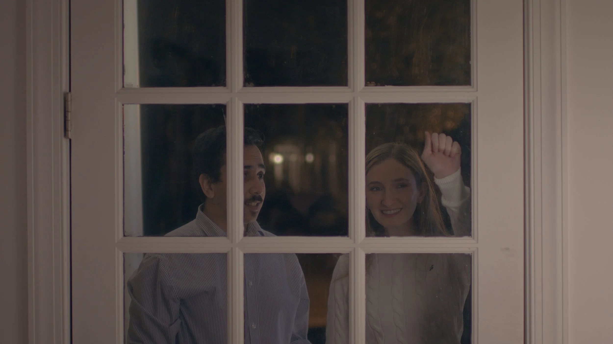 A man and woman are seen through a glass-paneled door, smiling and knocking, with internal lighting illuminating their faces and a dark outdoor background.