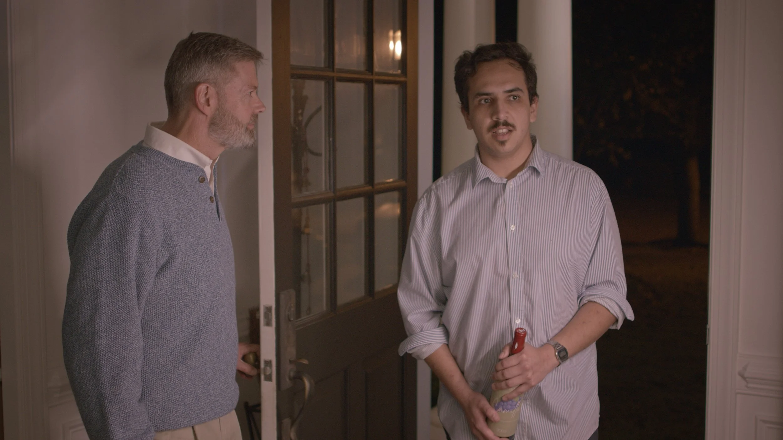 Two men standing at the doorway of a house, one holding a bottle and the other with his hand on the door handle, during nighttime.