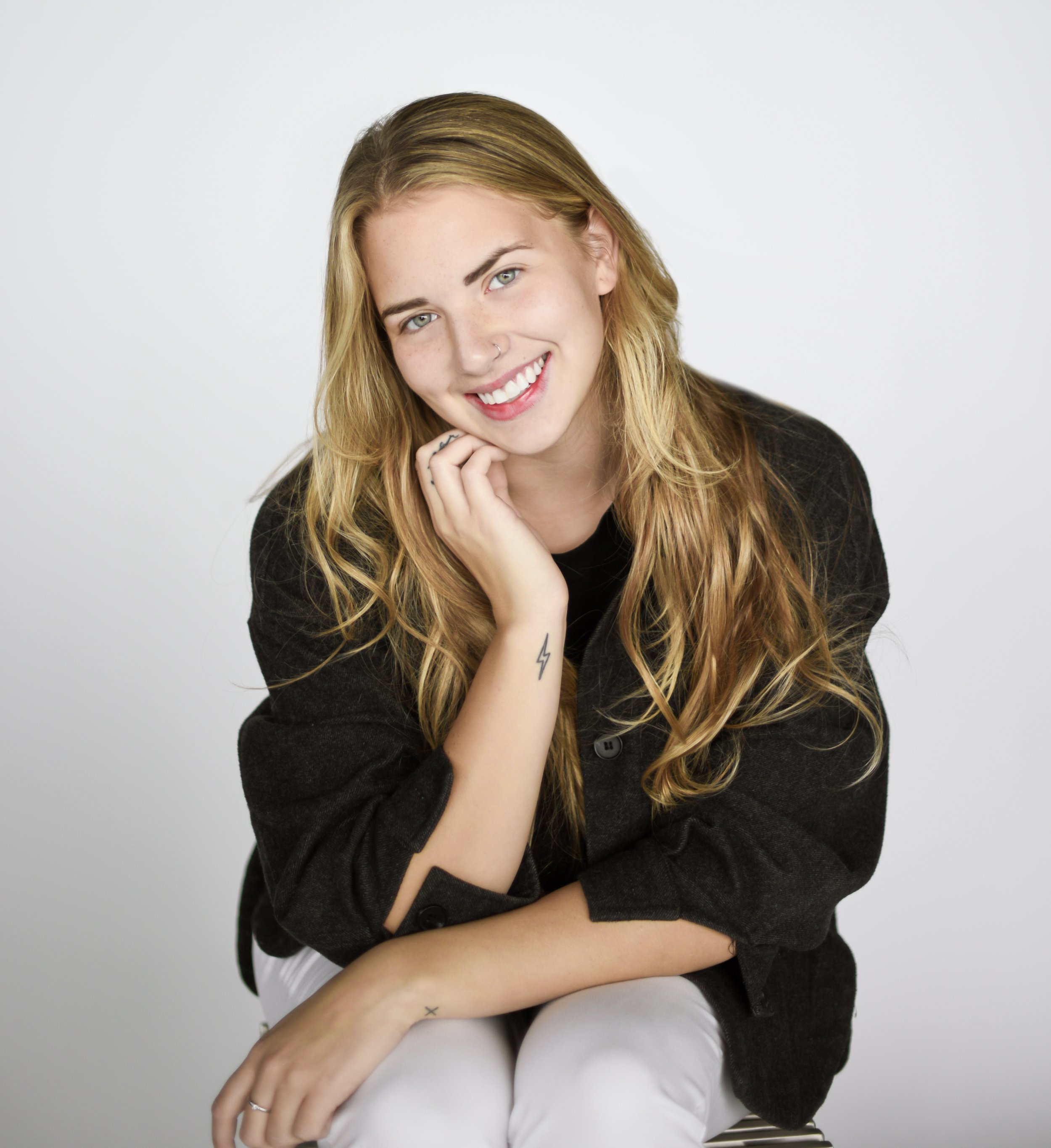 Carmen Halphen. A young woman with long blonde hair, blue eyes, and a bright smile, sitting against a plain white background, wearing a black jacket and white pants, with a small lightning tattoo on her wrist and a nose piercing.