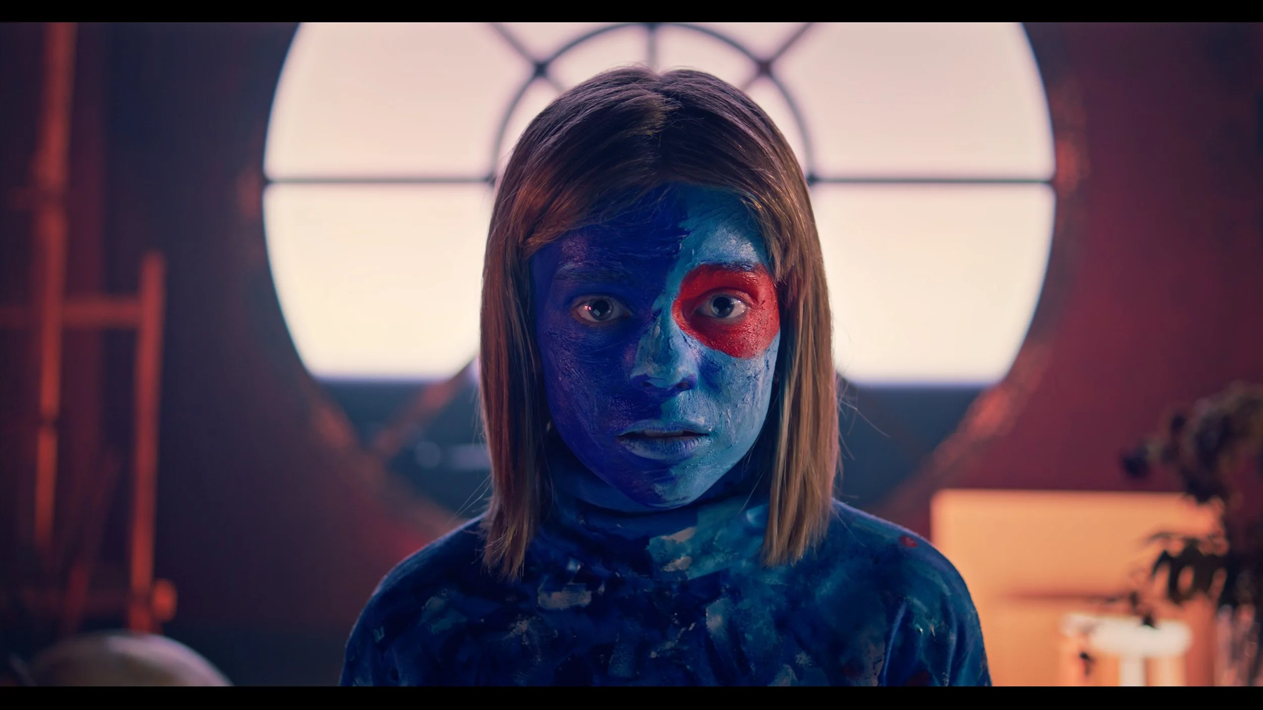 A young girl with face and upper body painted in blue, with red paint around her right eye, staring directly at the camera. A window with divided panes is visible in the background, inside a room with warm lighting.