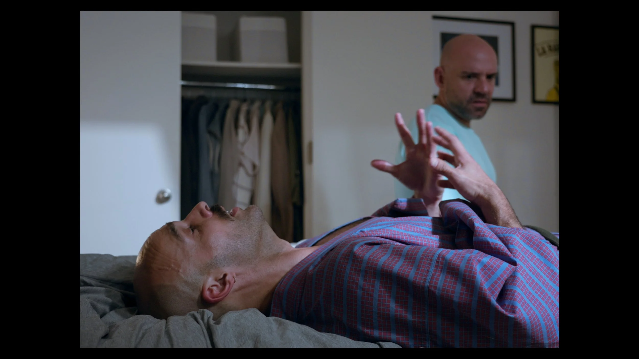 A man lying on a bed with eyes closed and hand raised, while another man sitting nearby looks at him with a serious expression, in a bedroom with an open closet behind them.