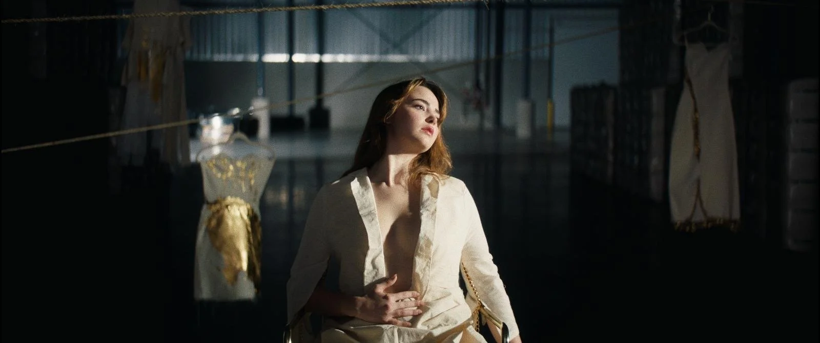 A woman with red hair and fair skin sitting on a chair in a dimly lit room, illuminated by a bright light, wearing a cream-colored dress, with shelves and cloths in the background.