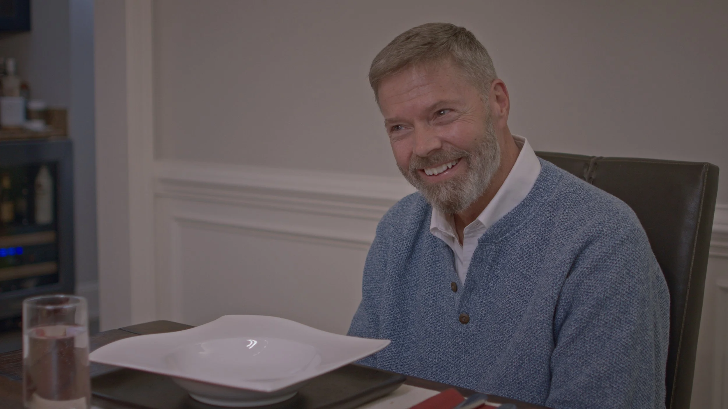 A smiling middle-aged man with a beard and short gray hair sitting at a dining table, wearing a white shirt and a blue sweater, with empty plates and a glass of water on the table.