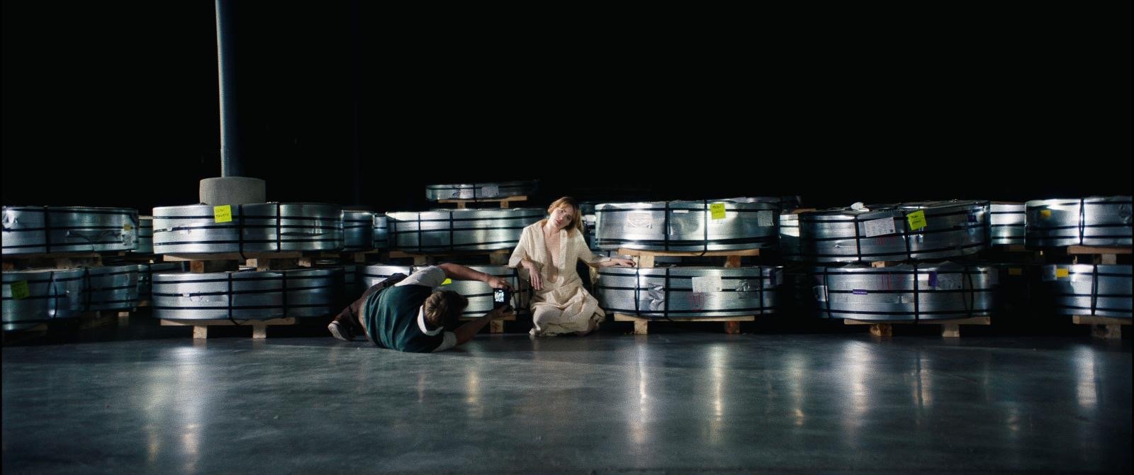 A woman in a light-colored dress reclines on the floor in front of a row of large metal objects stored on pallets in a dark industrial warehouse. A man in a dark vest and white shirt is kneeling and reaching towards her.