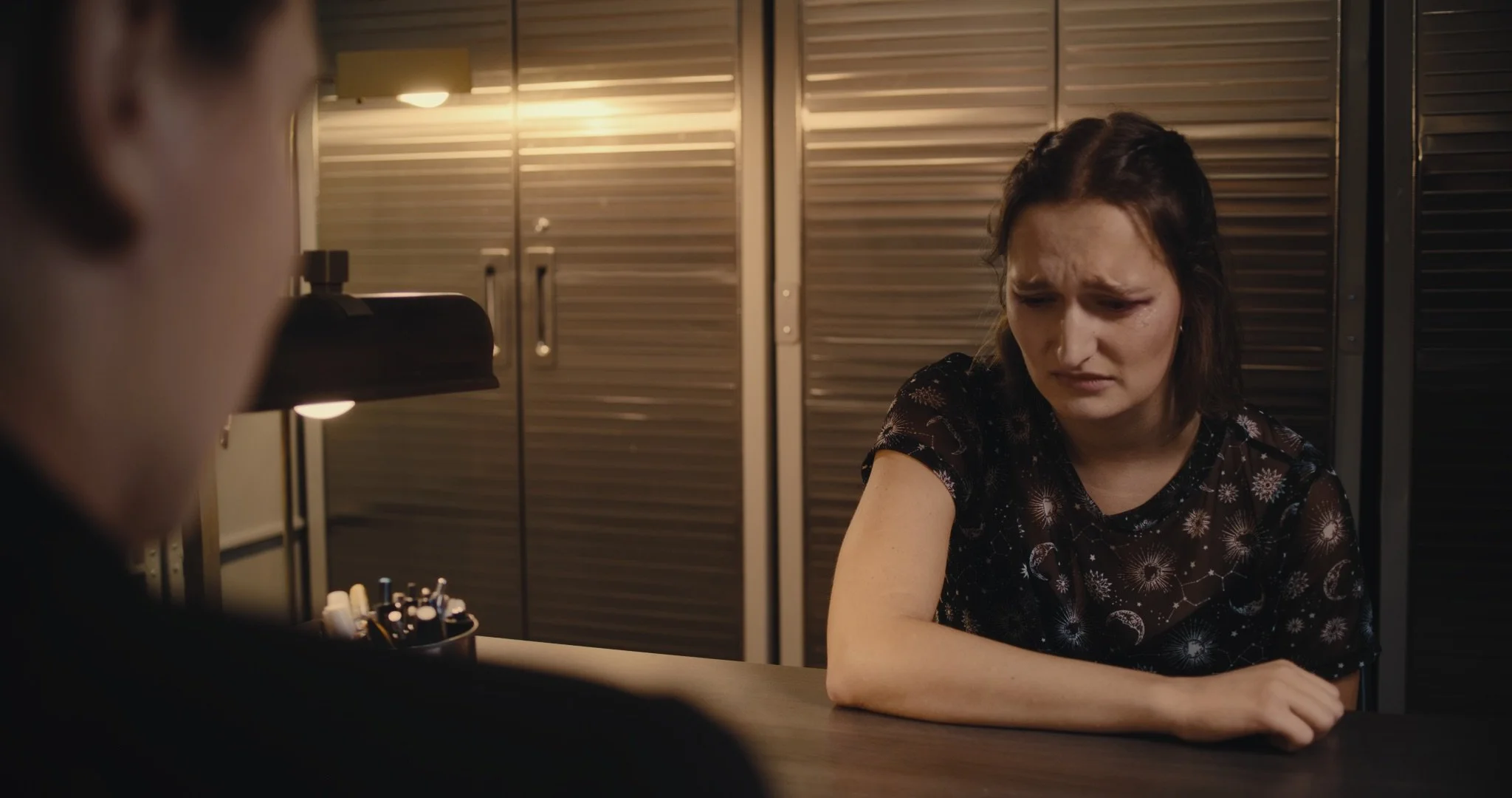 A woman sitting at a desk with a somber or upset expression, with a person blurred in the foreground, in a room with metal lockers in the background.