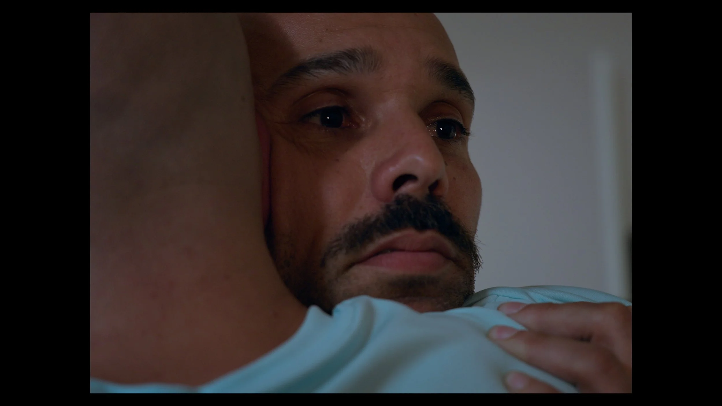 A man with a beard and mustache has a solemn, emotional expression while being hugged. The focus is on his face, showing a close-up of his eyes and lips, with another person hugging him from behind, their shoulder visible.