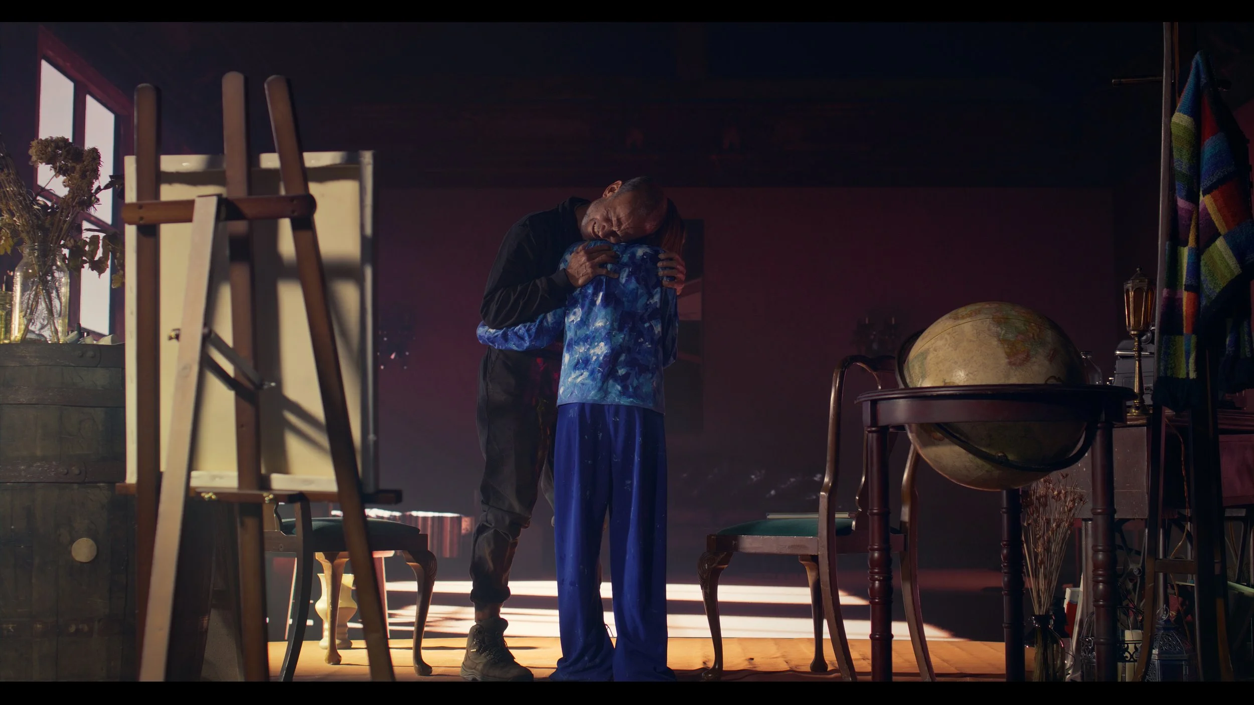 A man embraces a young girl in a dimly lit room filled with furniture and decorations, with a large globe on a table on the right.