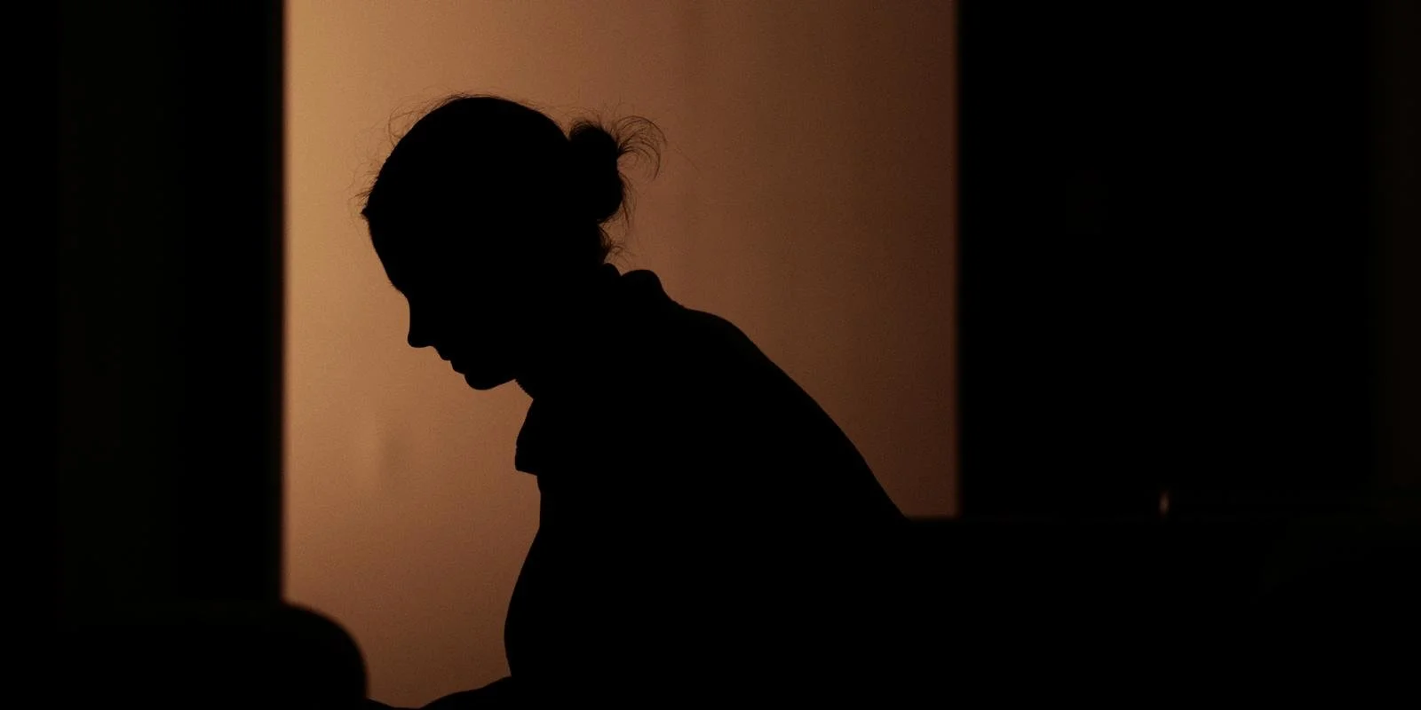 Silhouette of a person with their head bowed, sitting in a dimly lit room with warm lighting.