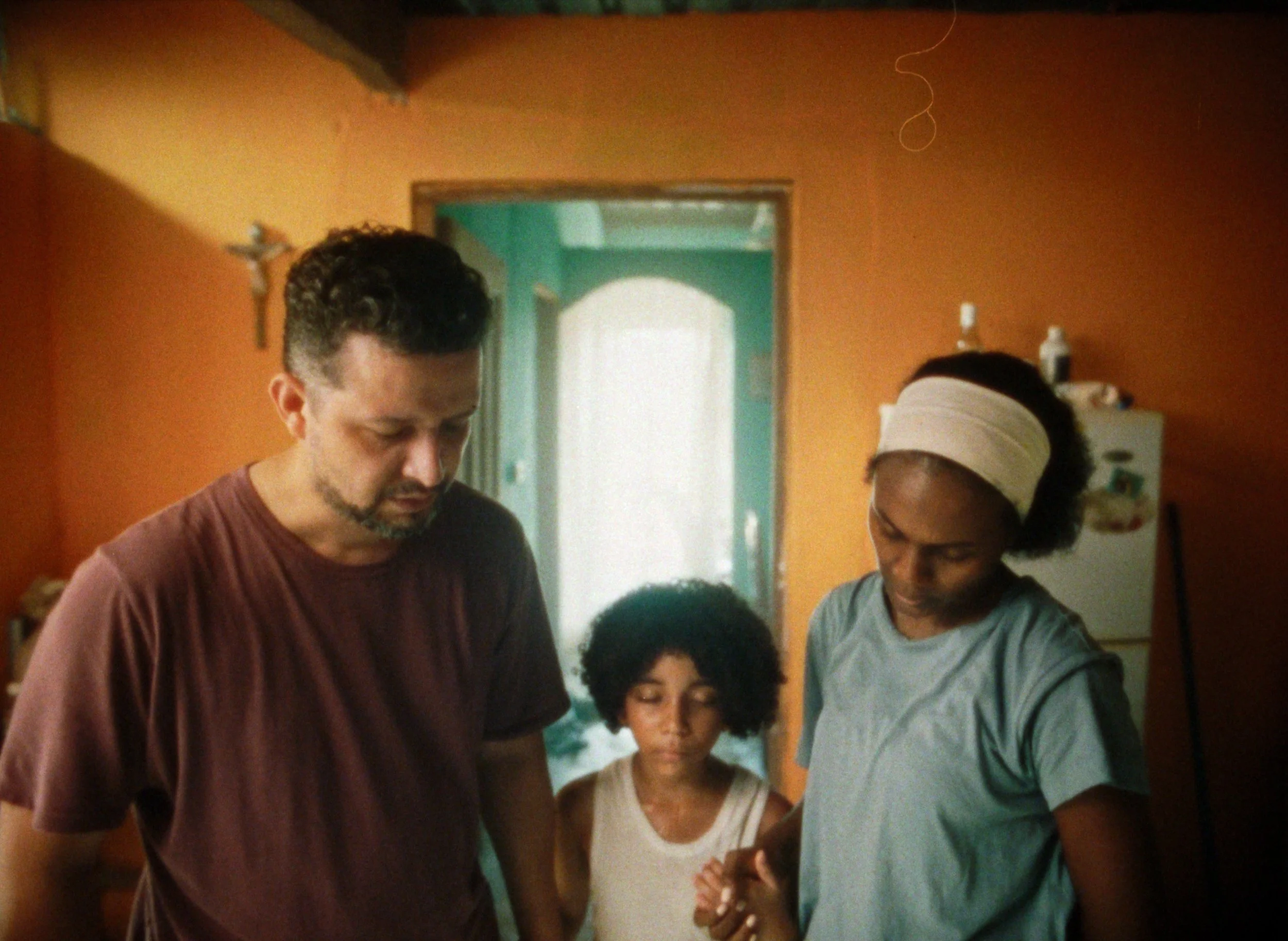 A man, woman, and young girl stand together in a kitchen with orange and blue walls, looking down in front of them.