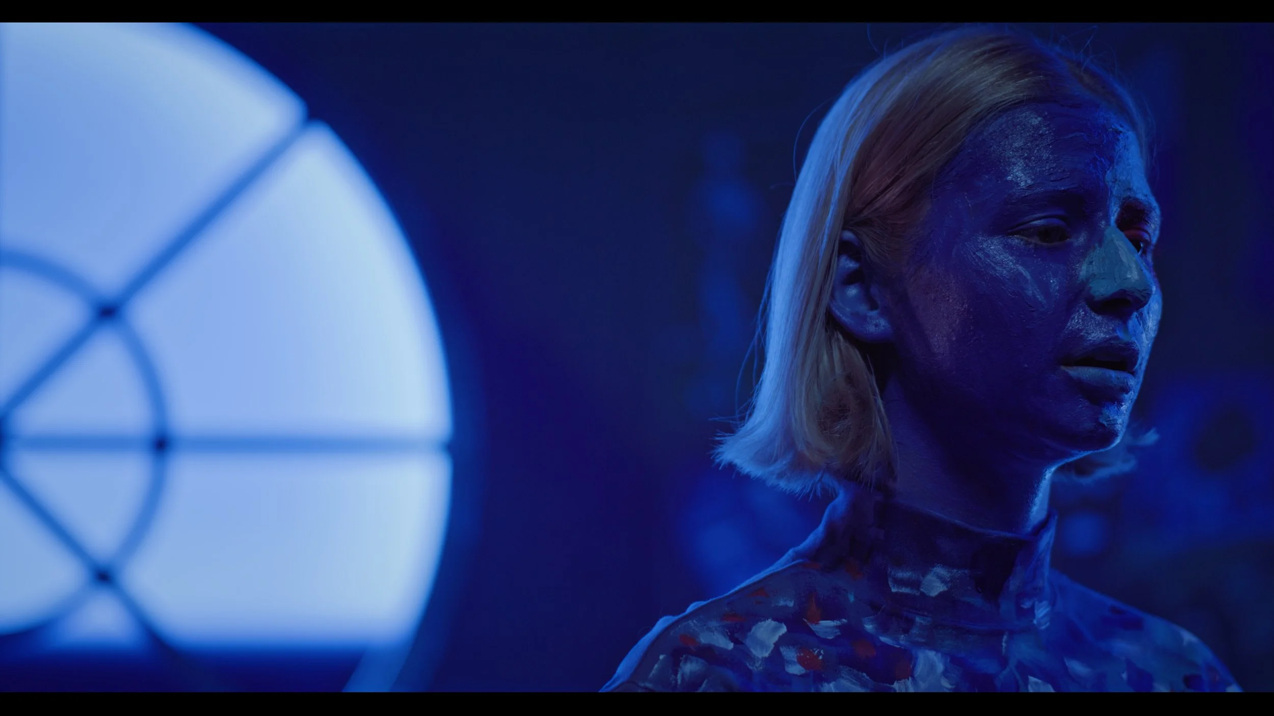 A woman with blonde hair covered in blue paint, looking to the right in a dimly lit environment with a blue stage light in the background.