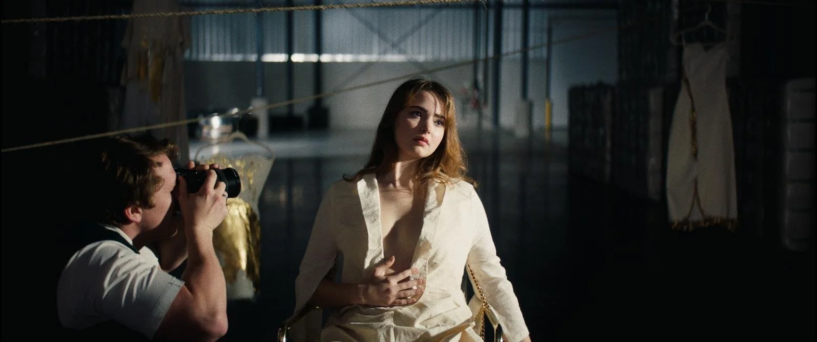 A woman with long, wavy hair sitting in a dimly lit room while a person with a camera takes her photo.