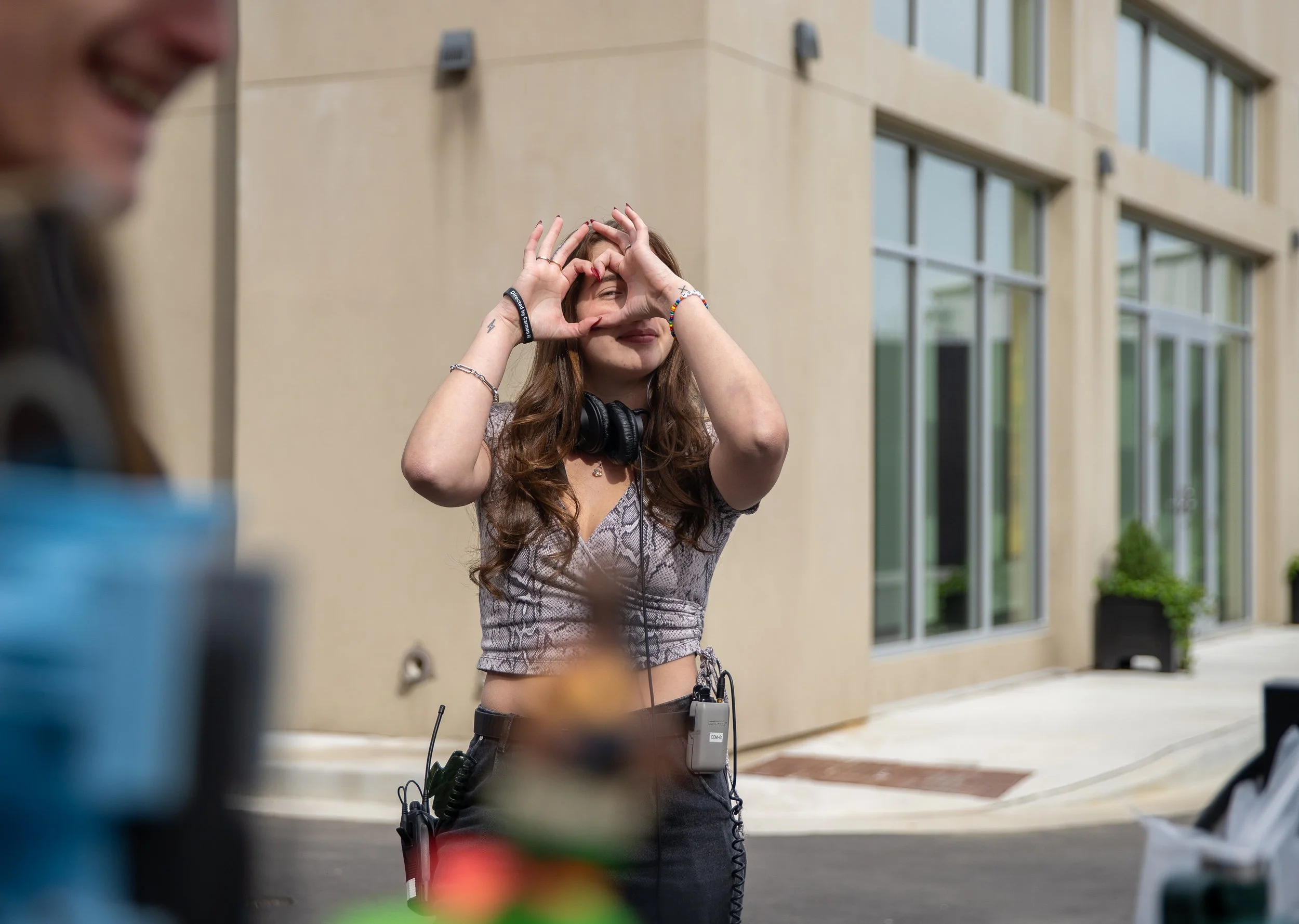 A young woman with long brown hair making a heart shape with her hands around her eyes, standing outside near a beige building with large windows.