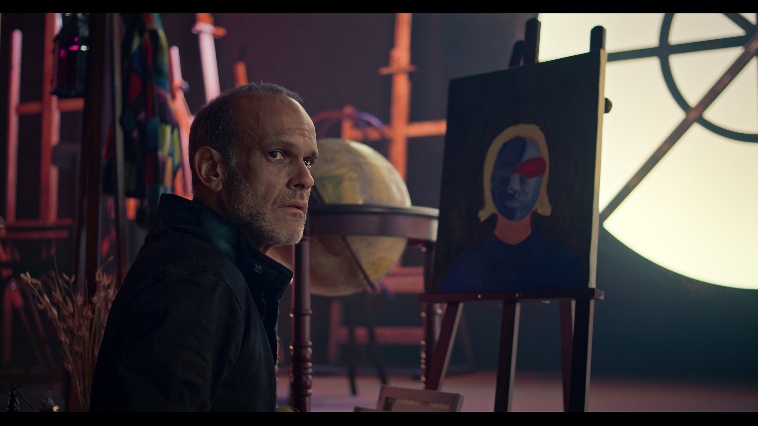 A middle-aged man with short hair and a beard, wearing a black shirt, looking over his shoulder in an art studio. Behind him are art supplies, a globe, and a painting of a woman with blonde hair, wearing a blue outfit and a red eye covering over one 