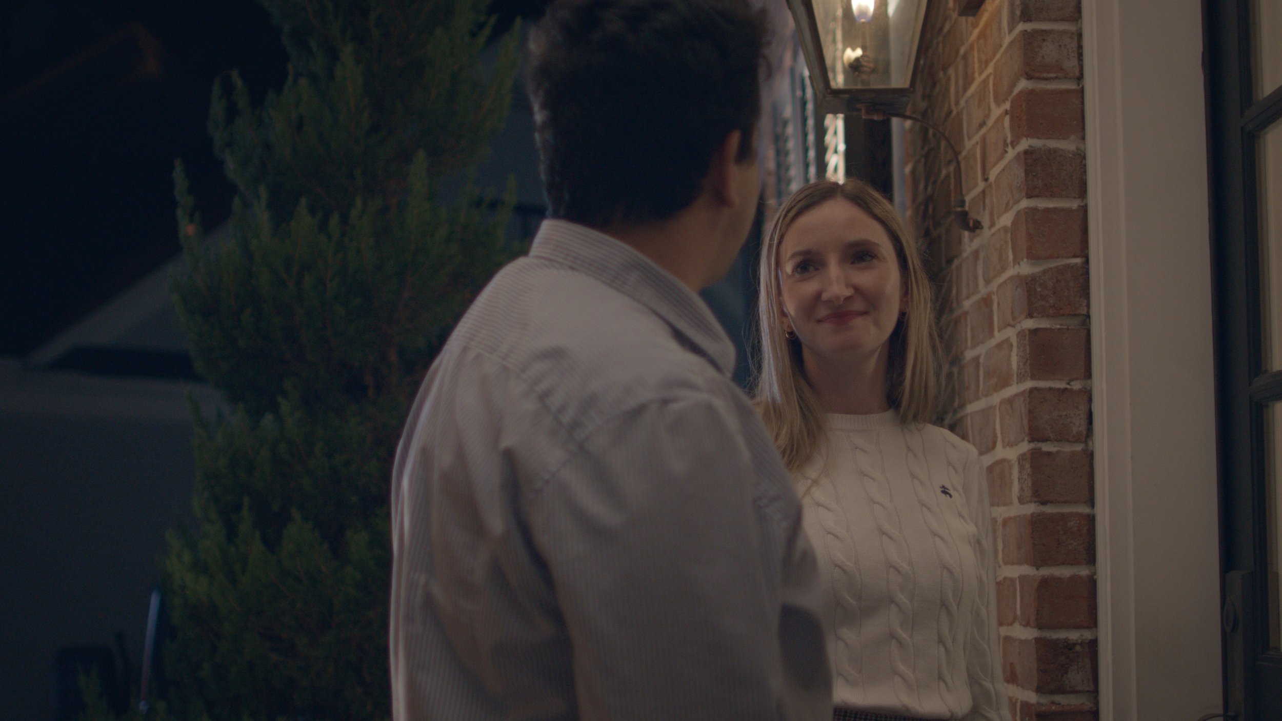 A woman with light blonde hair and a white cable-knit sweater smiling at a man with dark hair in a striped shirt, standing outside a brick house at night.