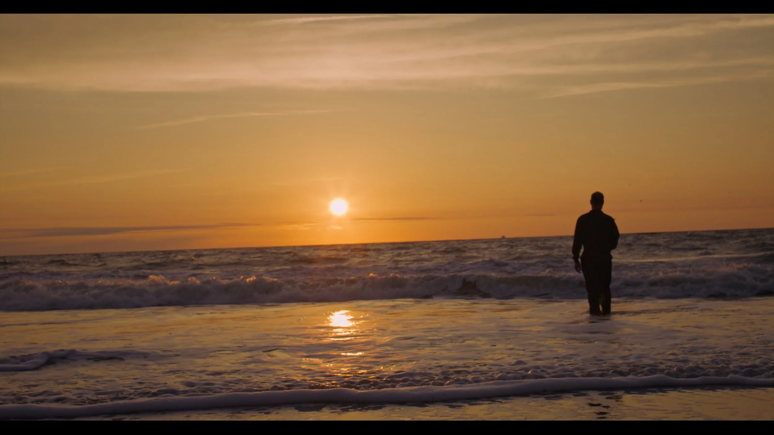 A person walking alone on the beach during sunset, with the sun low on the horizon and the sky painted in warm colors, gentle waves lapping at the shoreline.
