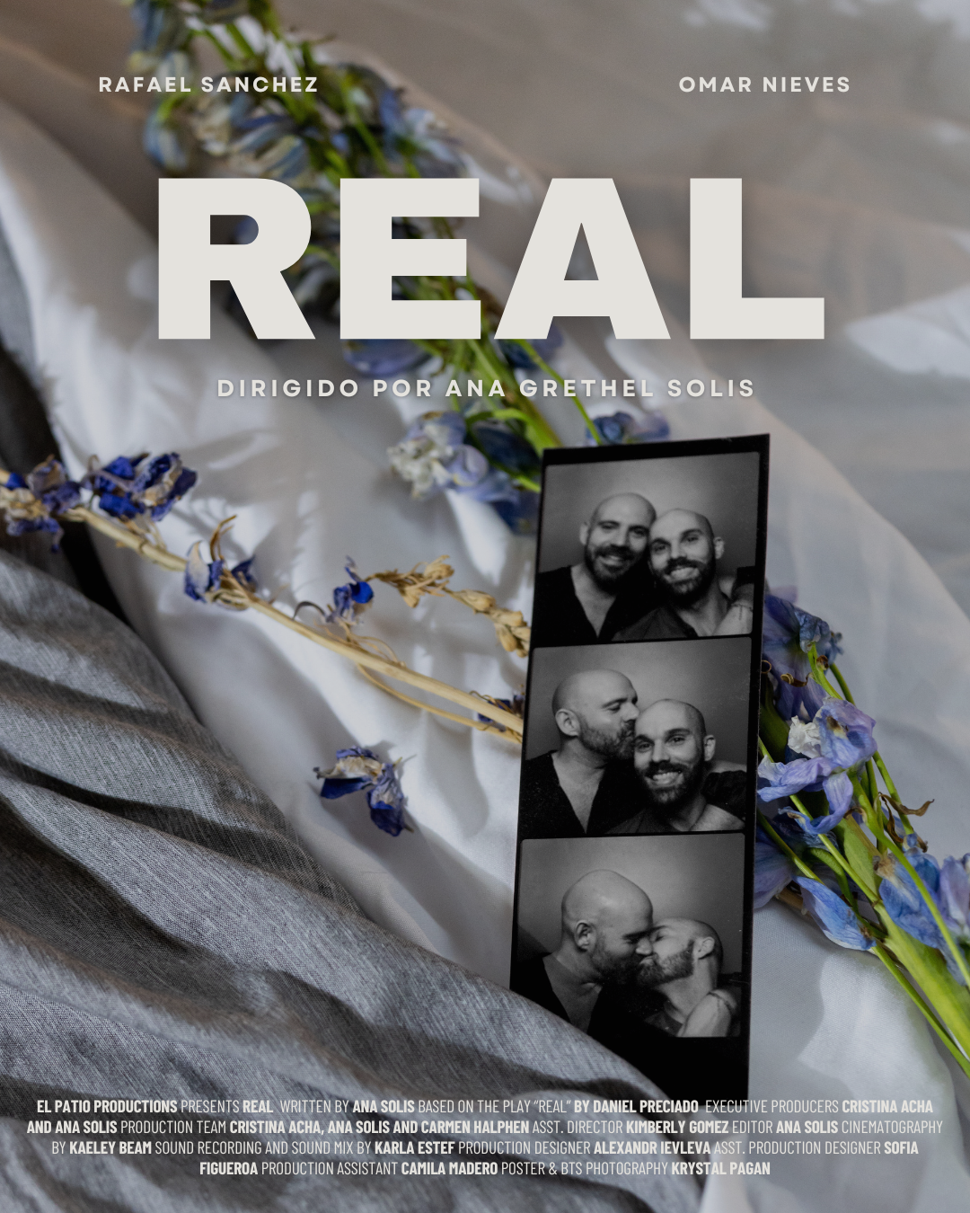 A promotional poster for the film 'Real' directed by Ana Solis produced by Carmen Halphen features, black-and-white photo booth image of two men sharing affectionate moments, laid on a surface with scattered blue and purple flowers and a white cloth.
