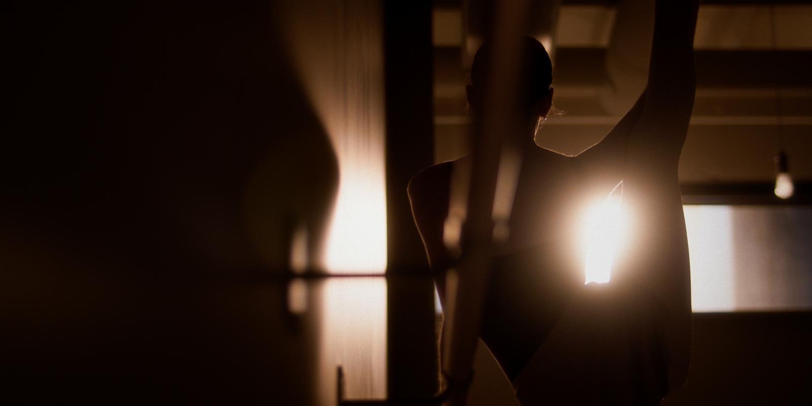 A woman is stretching in a dimly lit room with sunlight coming through a window behind her.