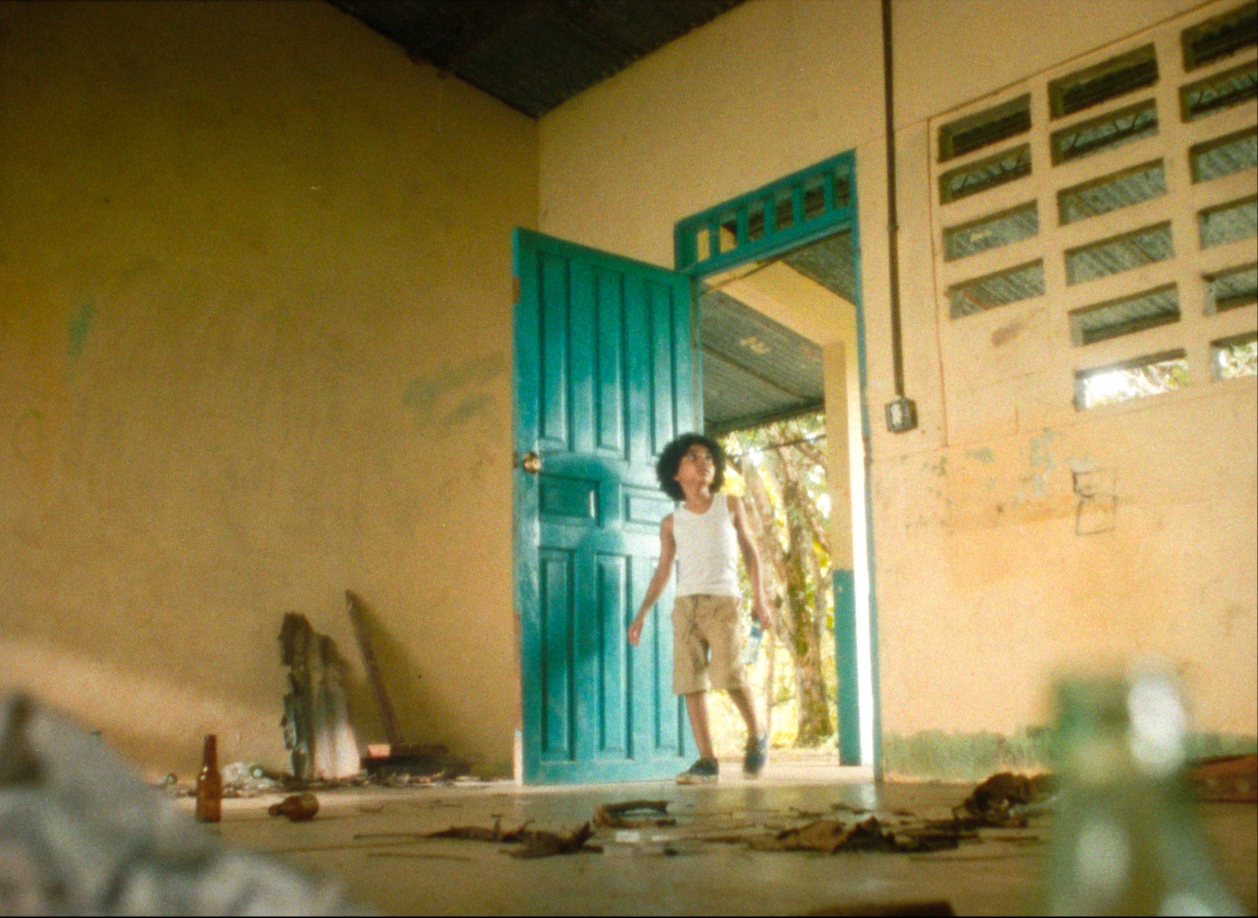 A young boy with curly black hair wearing a white sleeveless top and beige shorts, walking through an open turquoise door in a rustic, cluttered room with a concrete floor, peeling walls, and scattered objects.