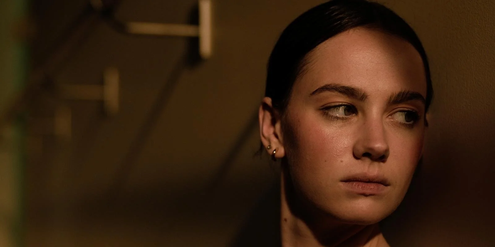 Close-up of a woman with dark hair and earrings, leaning against a wall, looking to the side with a contemplative expression.