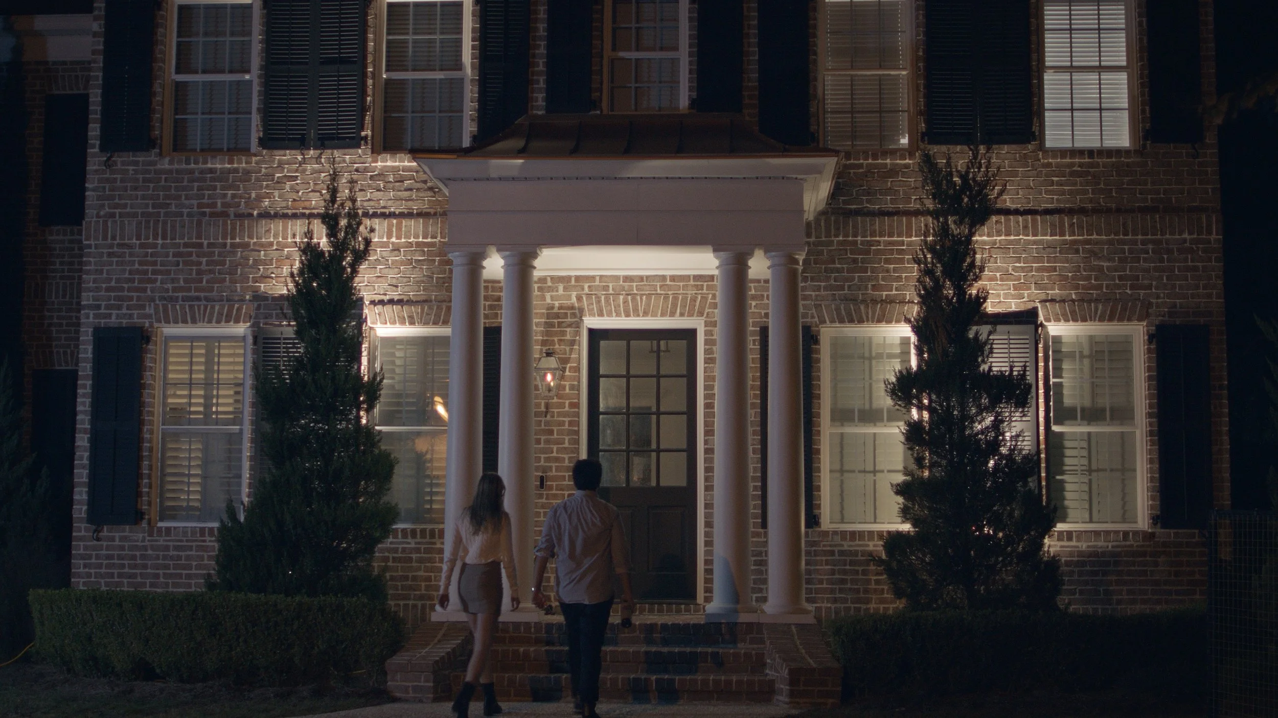 A couple walking up brick stairs to a porch of a brick house at night, with illuminated windows and tall bushes on either side.