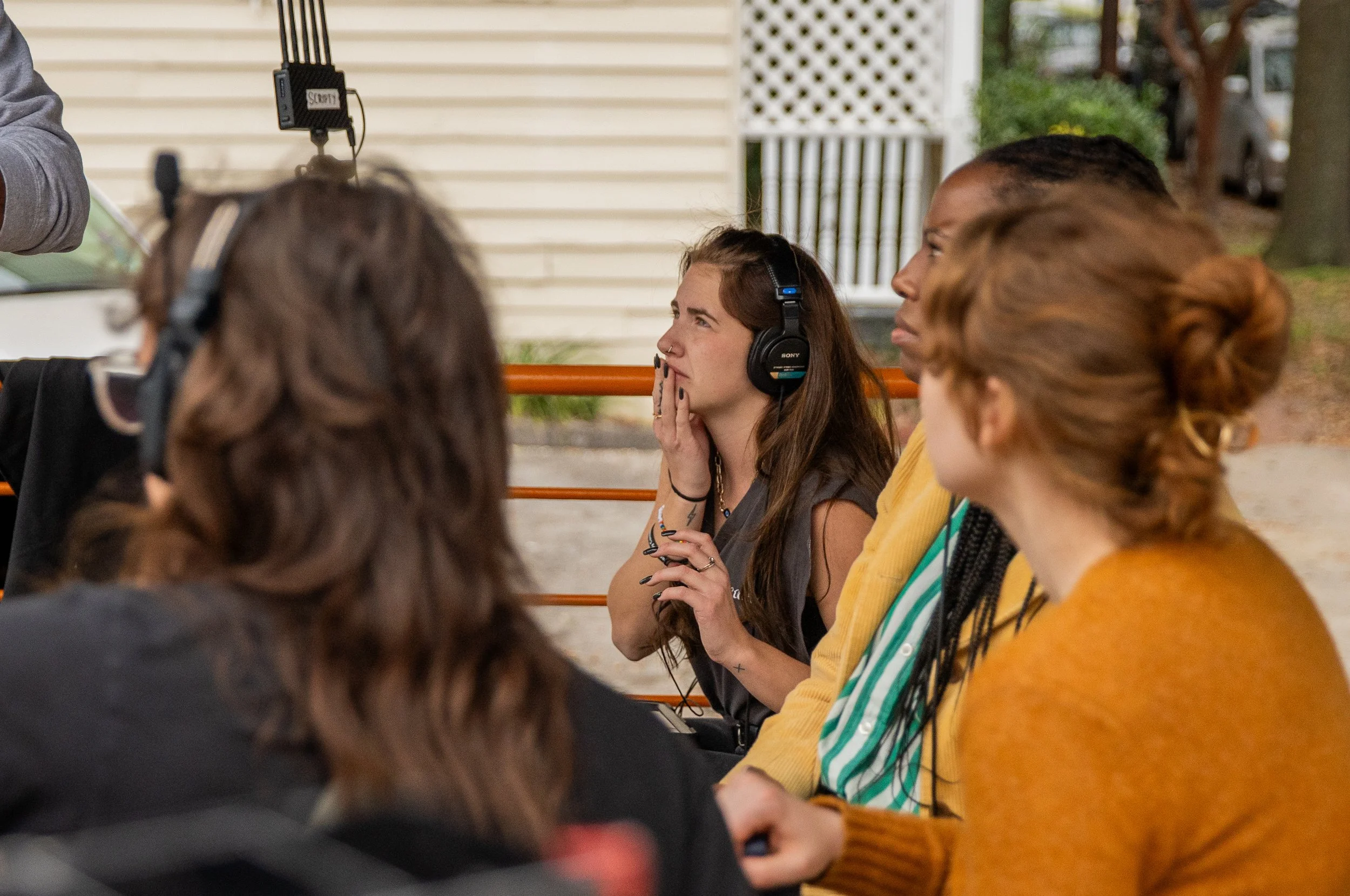A group of young women sitting on a bench, one wearing large headphones, outdoors in front of a house with a white lattice and trees.