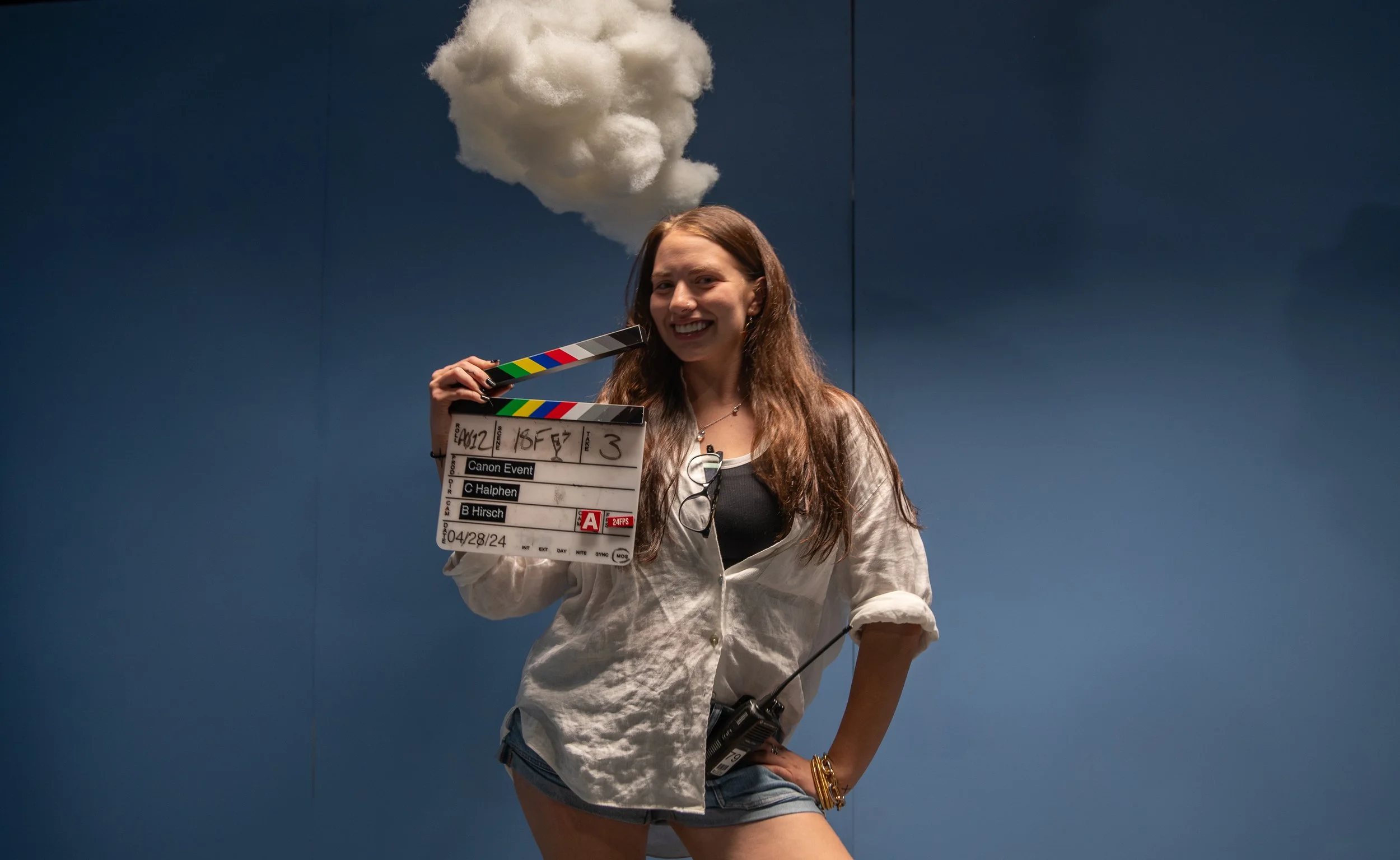 A woman holding a film slate stub with a cloud prop above her head, smiling, against a blue wall background.