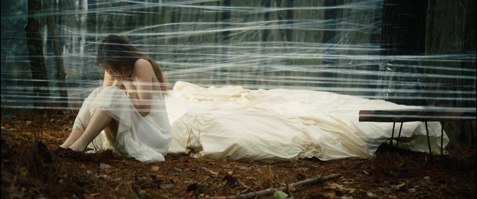 Woman dressed in white sitting on mossy ground with a white sheet or fabric covering a bed in the background, surrounded by trees, viewed through a translucent wrap encompassing her.