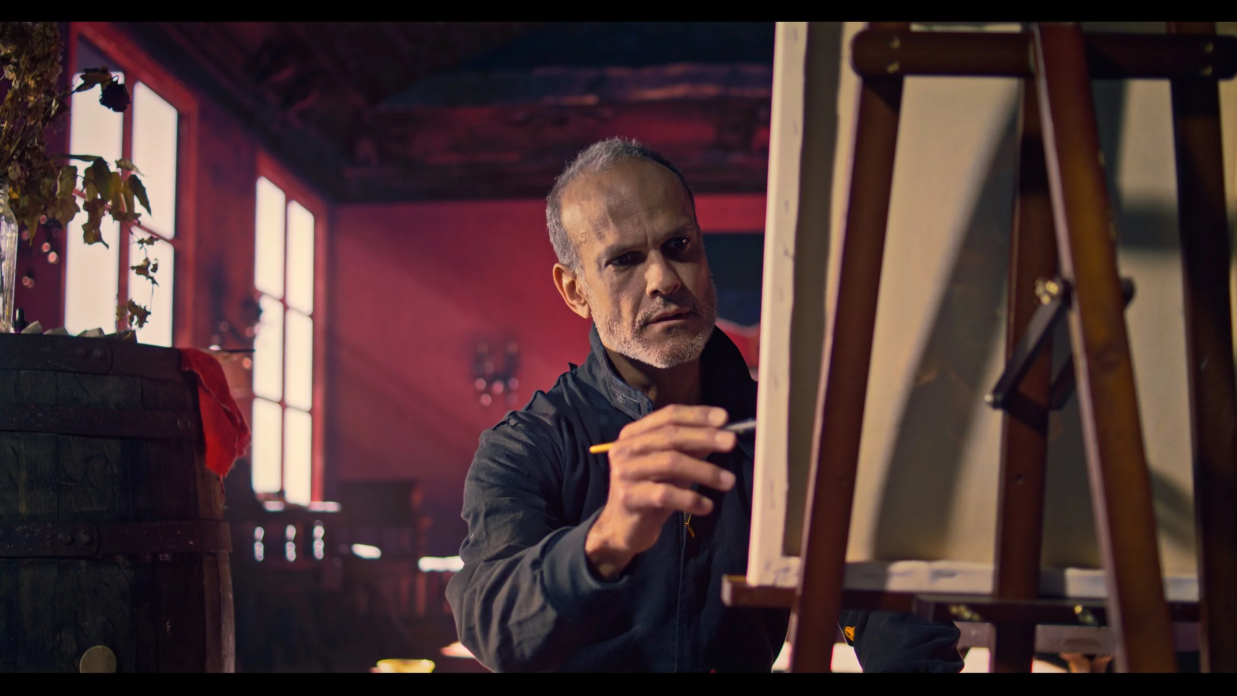 An older man with short, graying hair and a beard, wearing a black jacket, is painting on a canvas in a rustic, warmly-lit room with large windows and wooden beams.