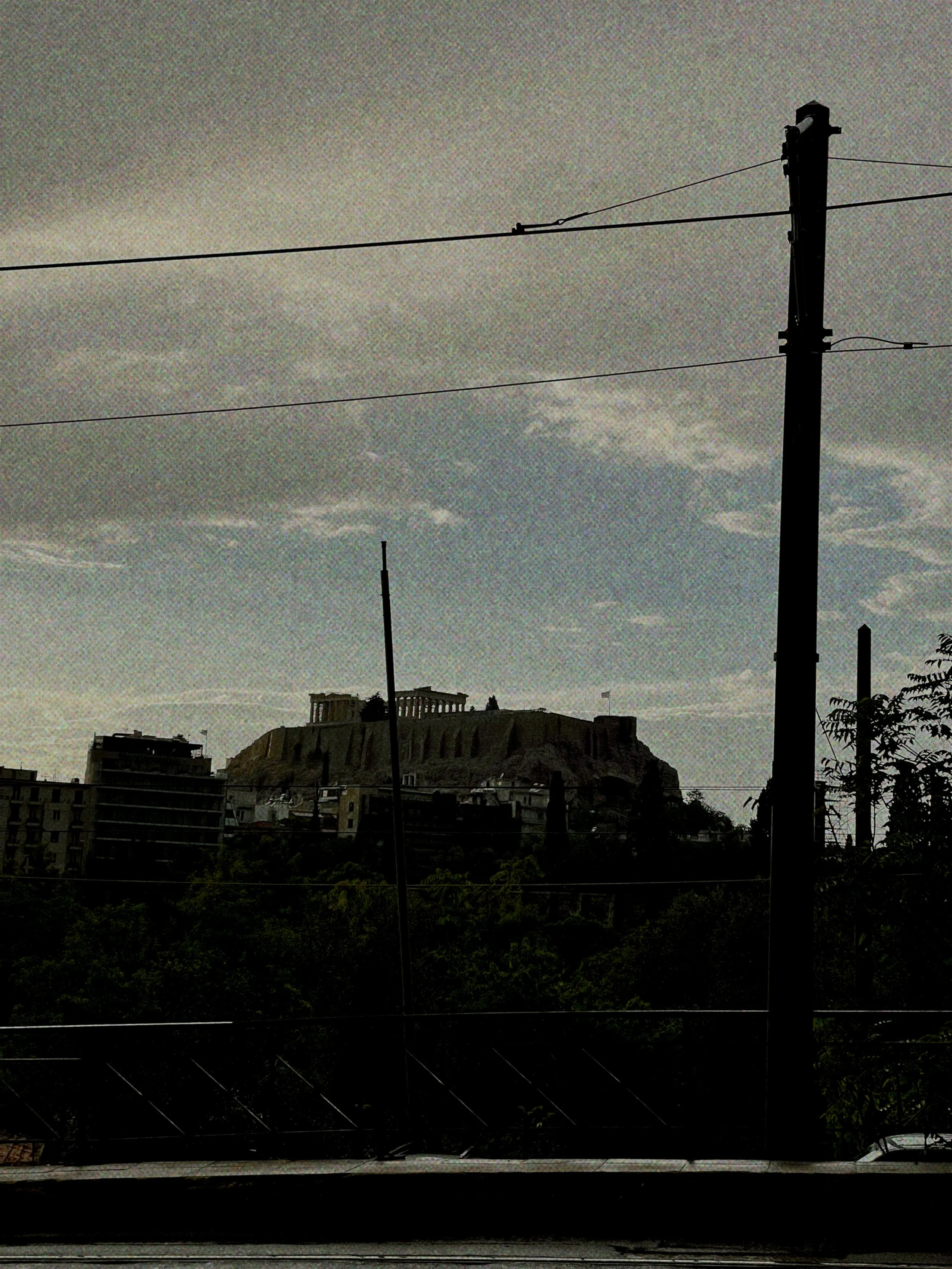 View of the Acropolis in Athens, Greece, seen from a distance with power lines and trees in the foreground.
