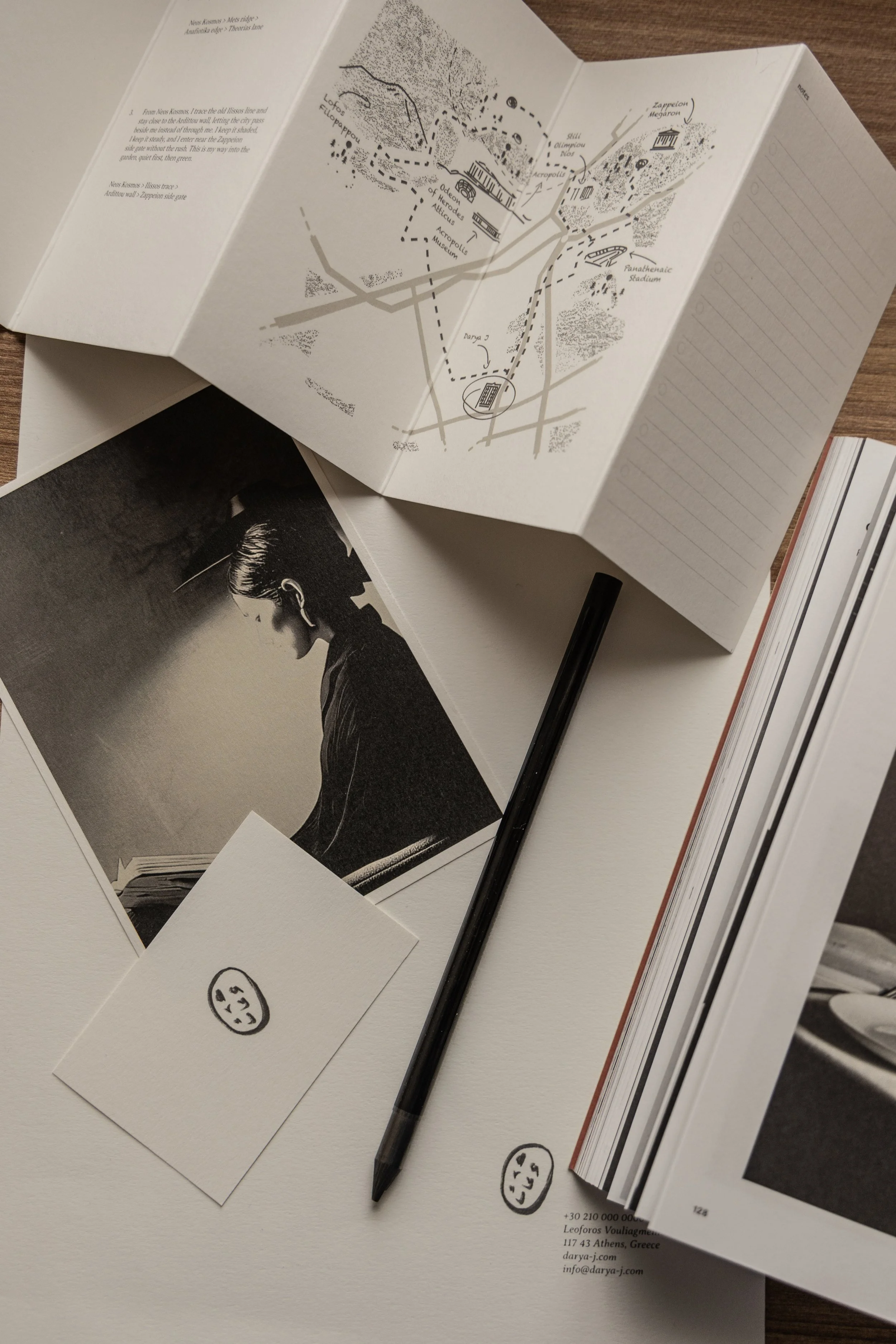 Open map, black and white photograph of a woman reading, a pen, sticky note with a smiley face, and a large open book on a wooden surface.