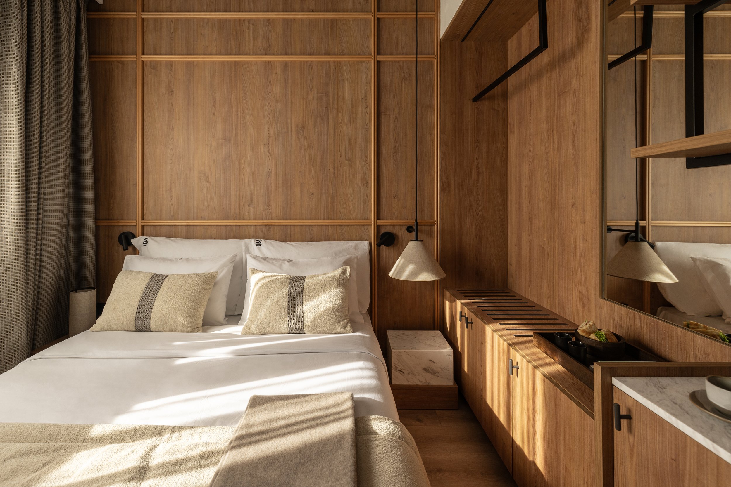 A cozy bedroom with wooden wall paneling, a bed with white sheets and beige pillows, a bedside table with a beige lamp, and a mirror on the right side.