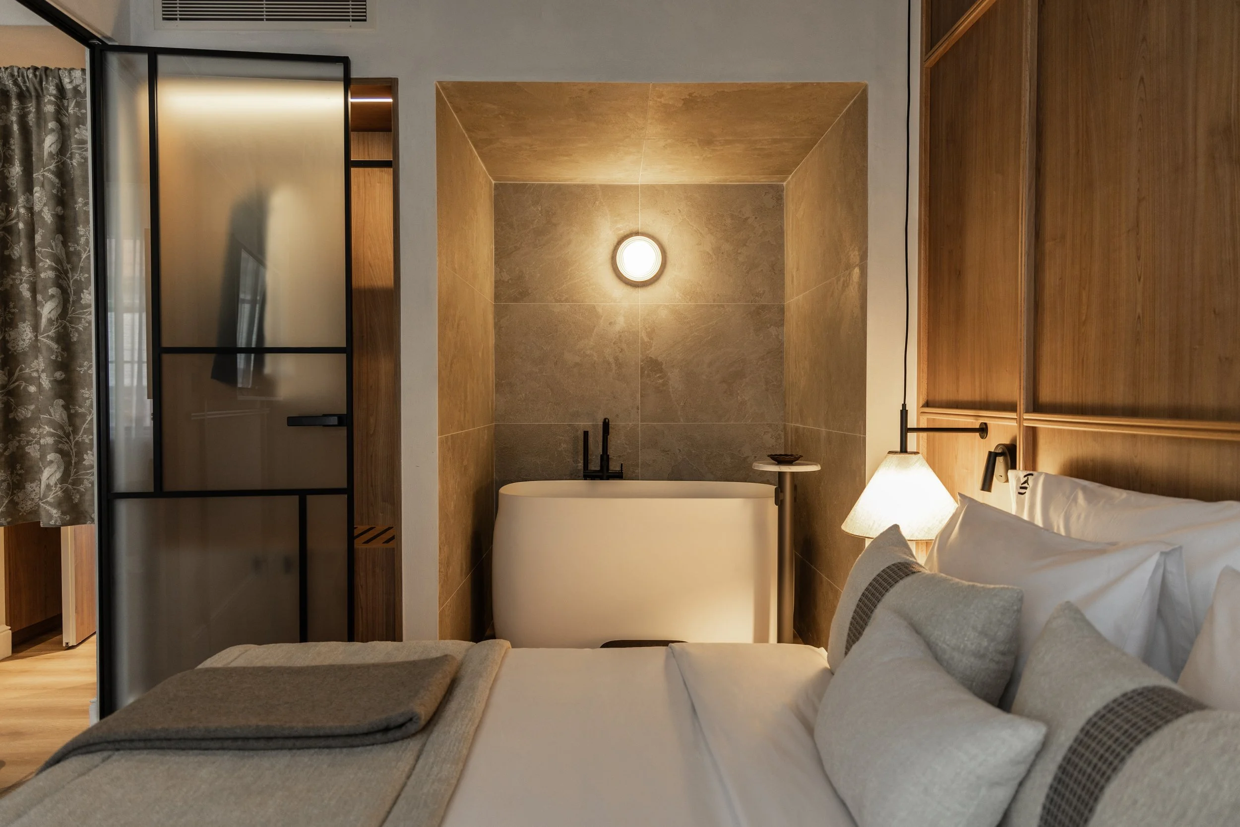 Hotel bedroom with a white bed and a standalone bathtub behind it, against a grey stone wall with a circular wall lamp, and part of a wooden wardrobe and sitting area in the background.