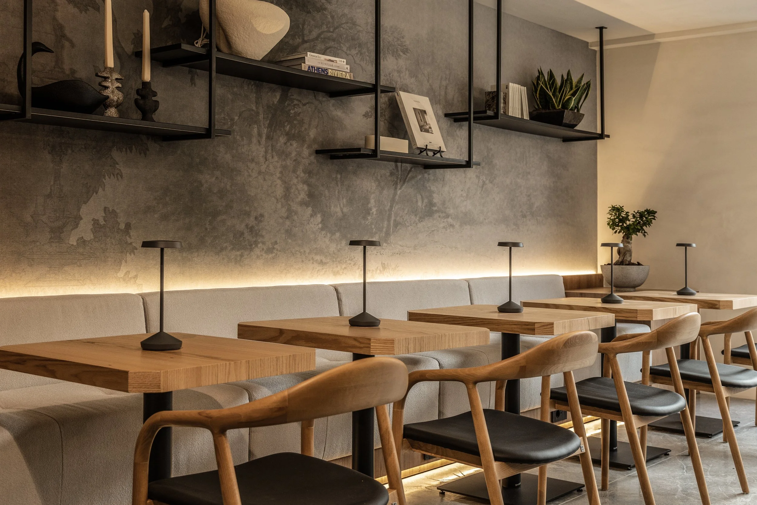 Interior of a modern restaurant with wooden tables, black chairs, beige upholstered bench seating, and decorative wall shelving with books and plants, illuminated by soft lighting.