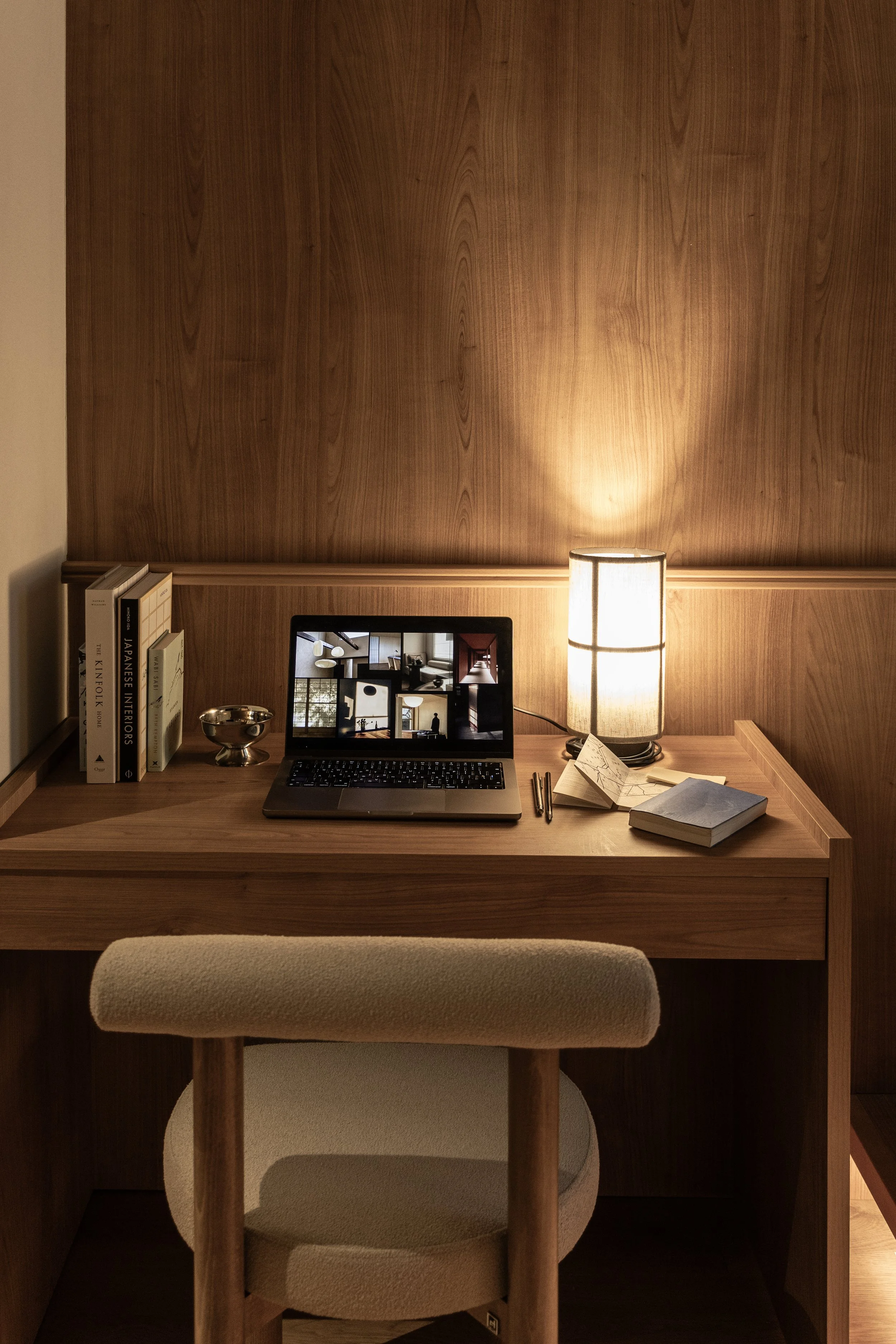 A cozy workspace with a wooden desk, a laptop displaying a gallery of images, a small stack of books, a lamp, pens, and notebooks, all set against a wooden panel wall.