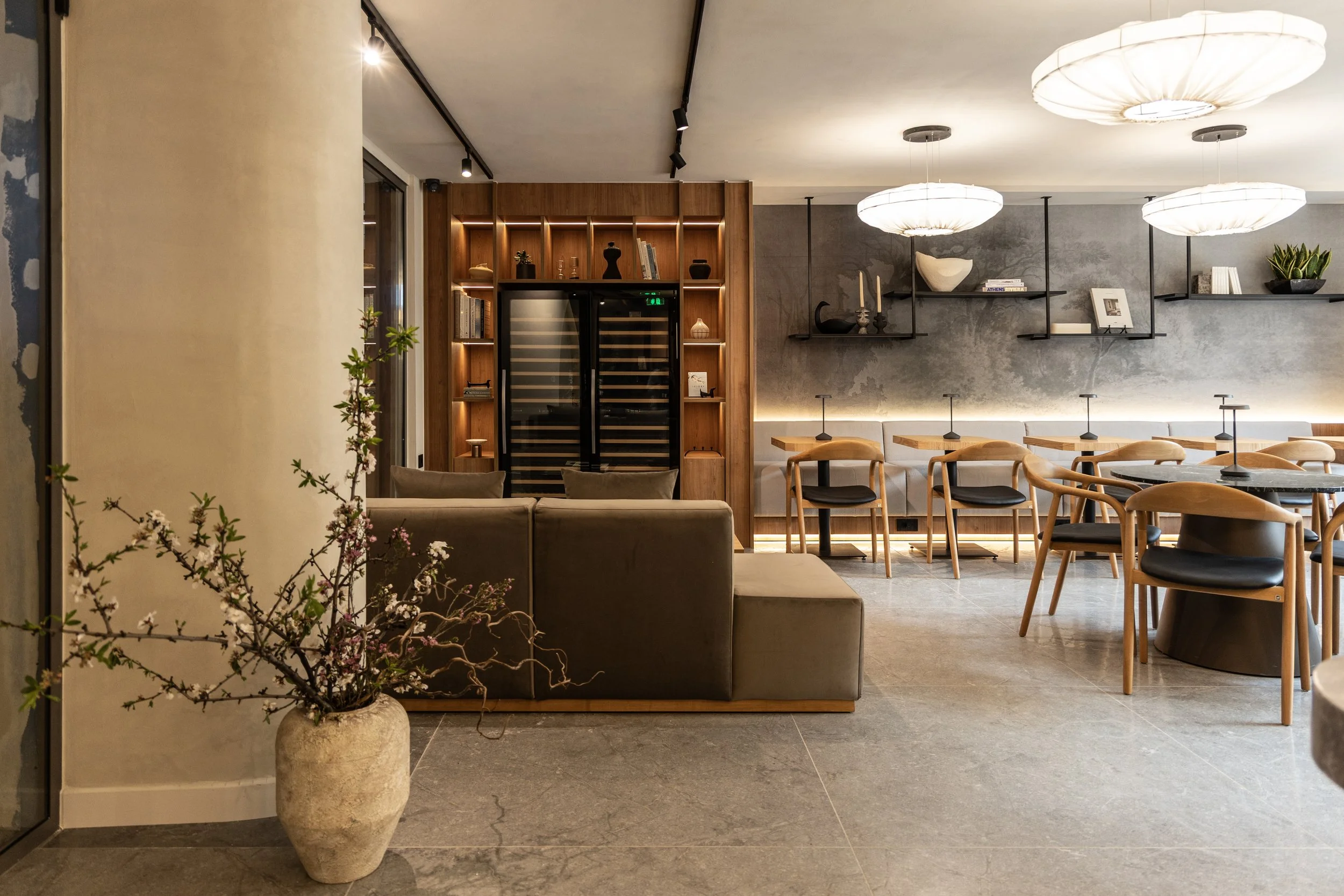 Modern interior of a lounge or restaurant with a potted plant in the foreground, beige sofa, wooden chairs and tables, a wall-mounted shelf with decorative items, and a wine fridge in the background, lit with warm, ambient lighting.