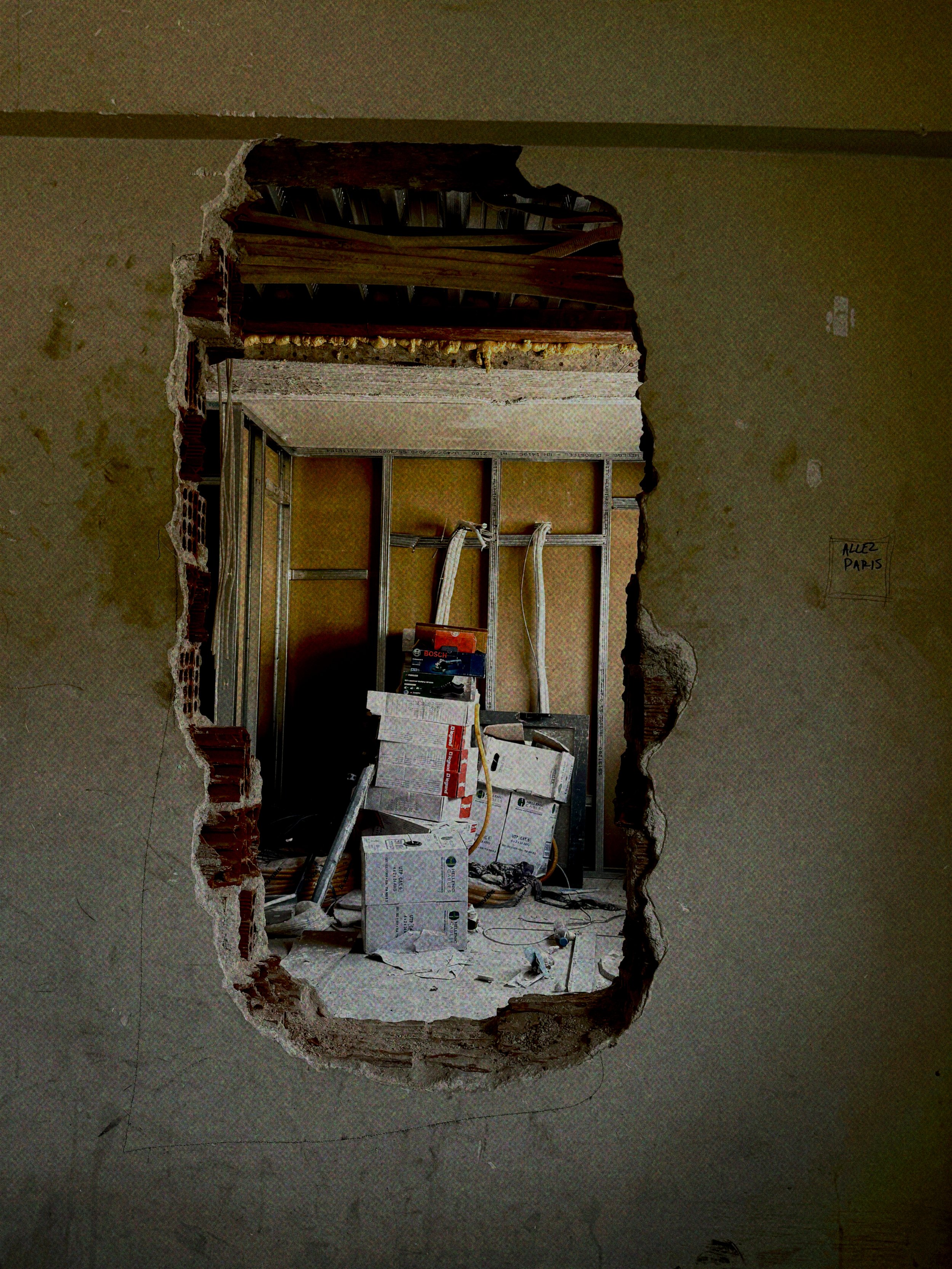 Interior view of a room under renovation, seen through a large irregular hole in a wall. The room contains construction tools, boxes, and exposed framing on the walls and ceiling.