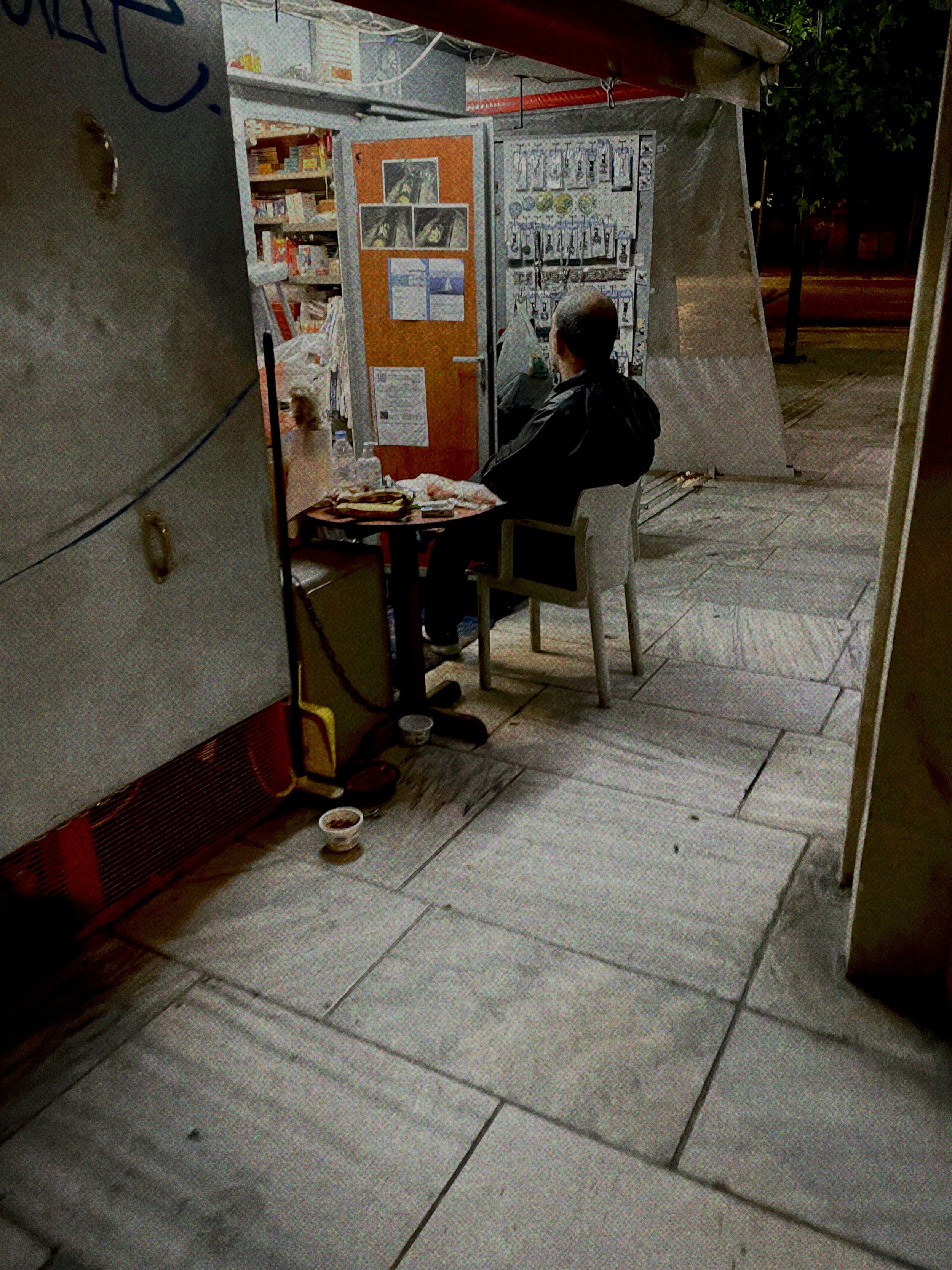 A man sitting at a small table outside a store at night, with various objects and posters on the wall behind him and some bowls on the ground nearby.