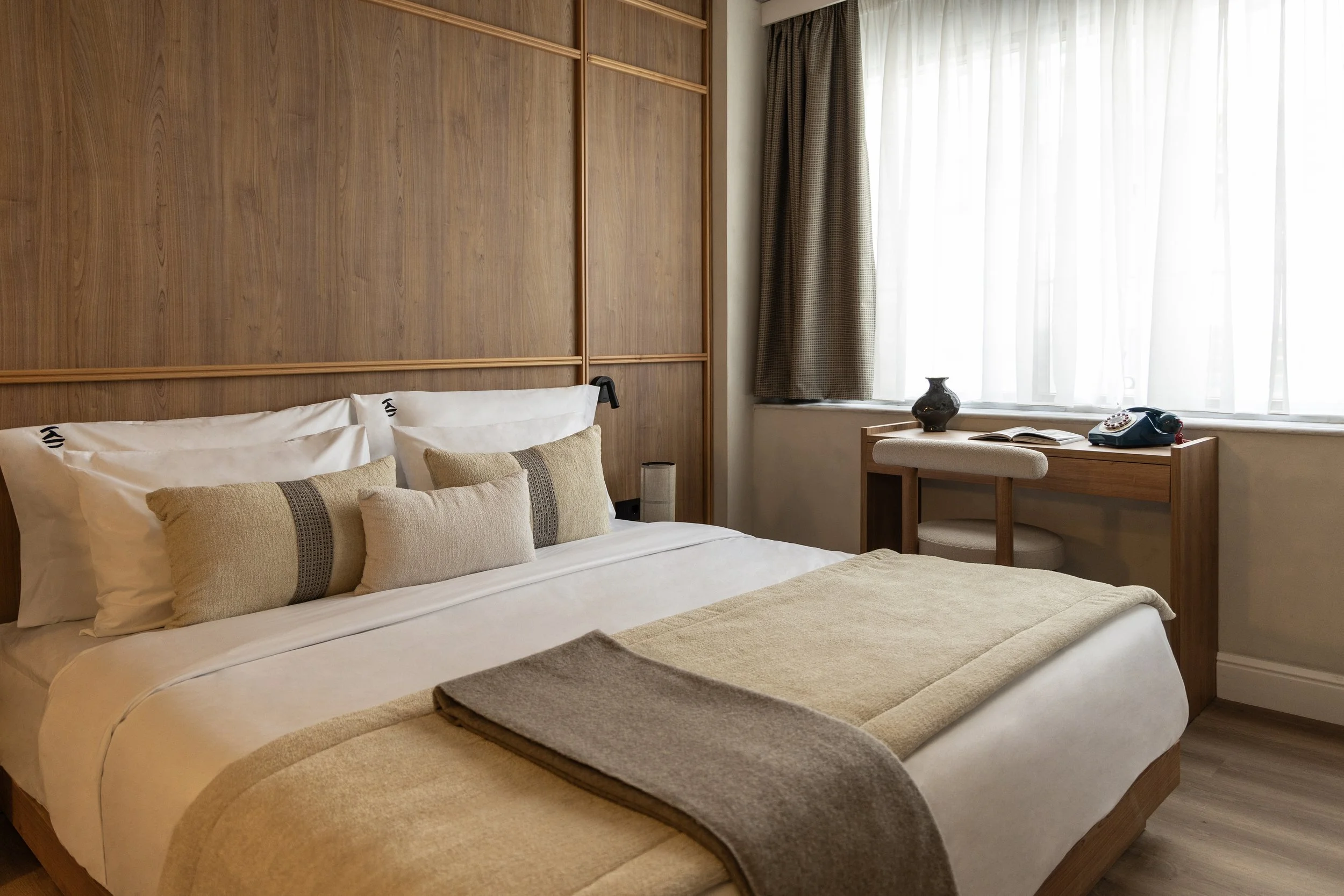 A neatly made hotel bed with pillows and a beige bedspread, next to a wooden headboard wall, with a window with curtains, a table with a vase, an open book, and a telephone on it.